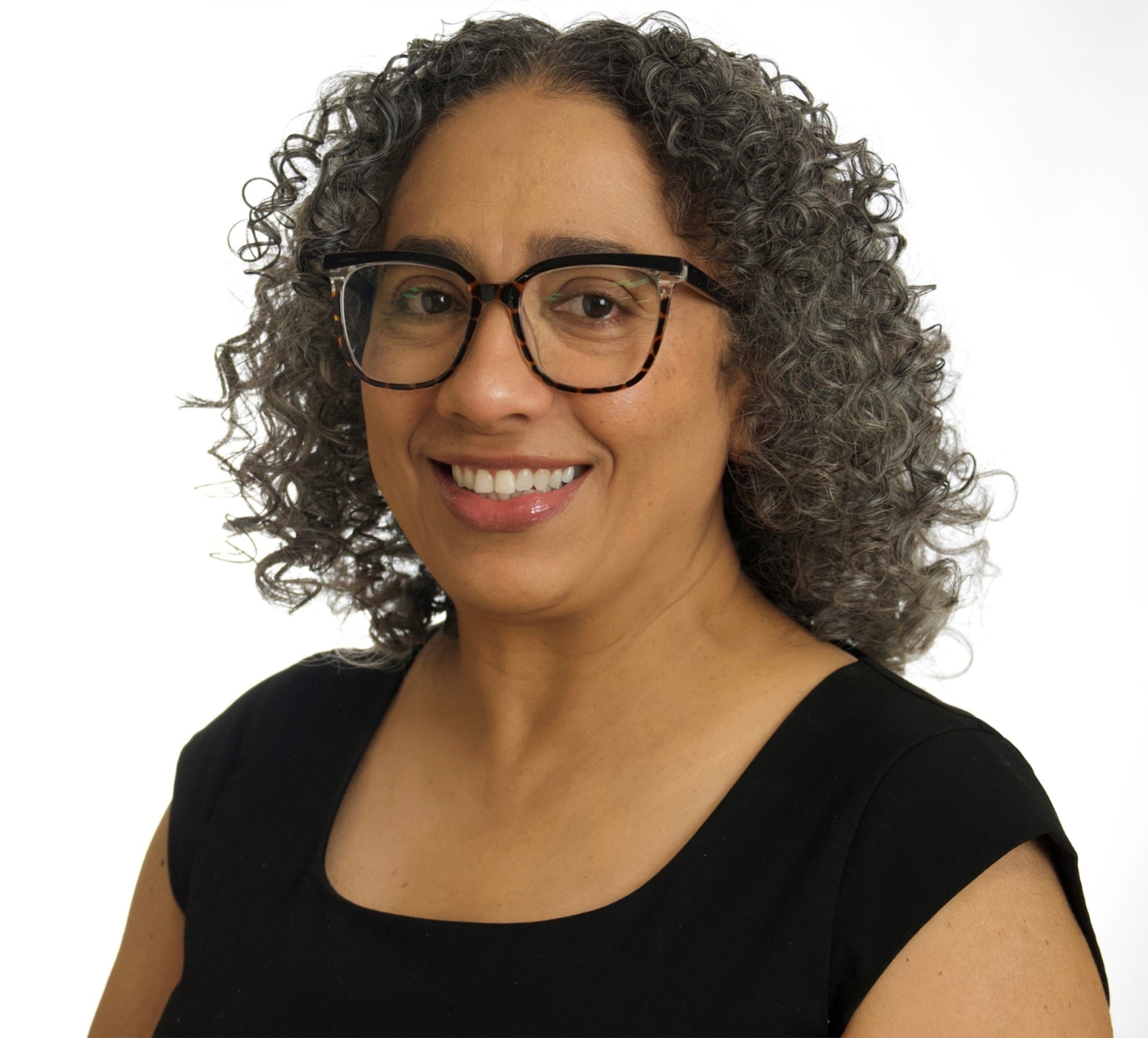 A middle-aged woman with curly gray hair and glasses, smiling, wearing a black sleeveless top against a white background.