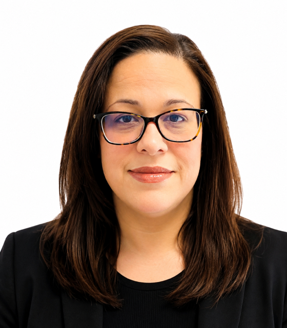 A woman with shoulder-length brown hair, wearing glasses with tortoiseshell frames, a beige sweater, and lipstick, looking at the camera against a white background.
