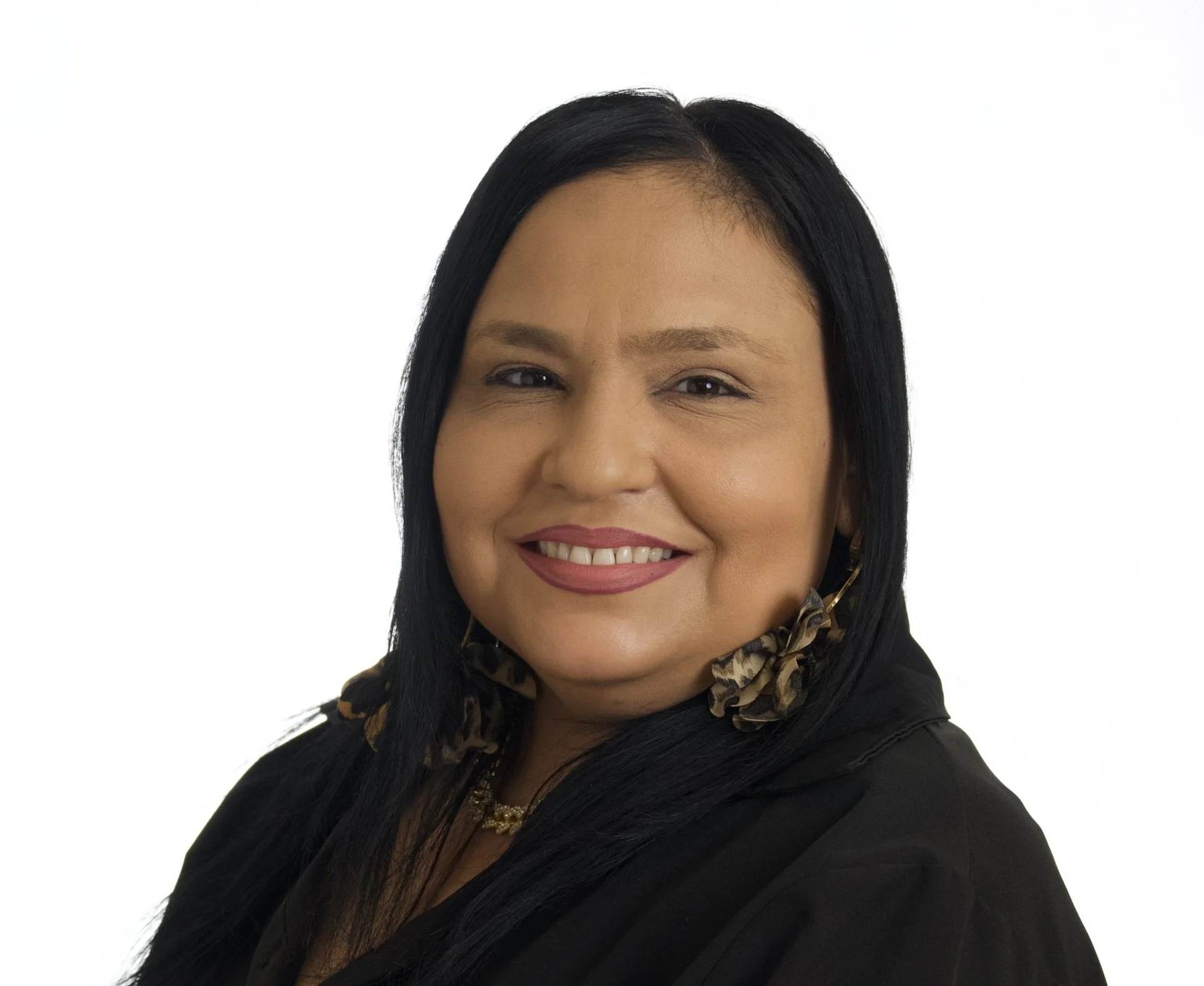 A woman with long black hair, wearing large leopard print earrings, a black top, and a gold necklace, smiling at the camera against a white background.