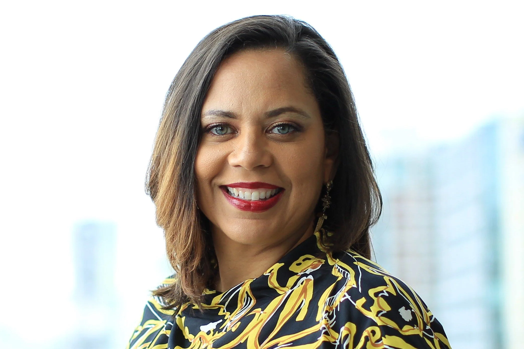 Portrait of Denise Ransom smiling, with shoulder-length brown hair, red lipstick, and a black and gold patterned top.