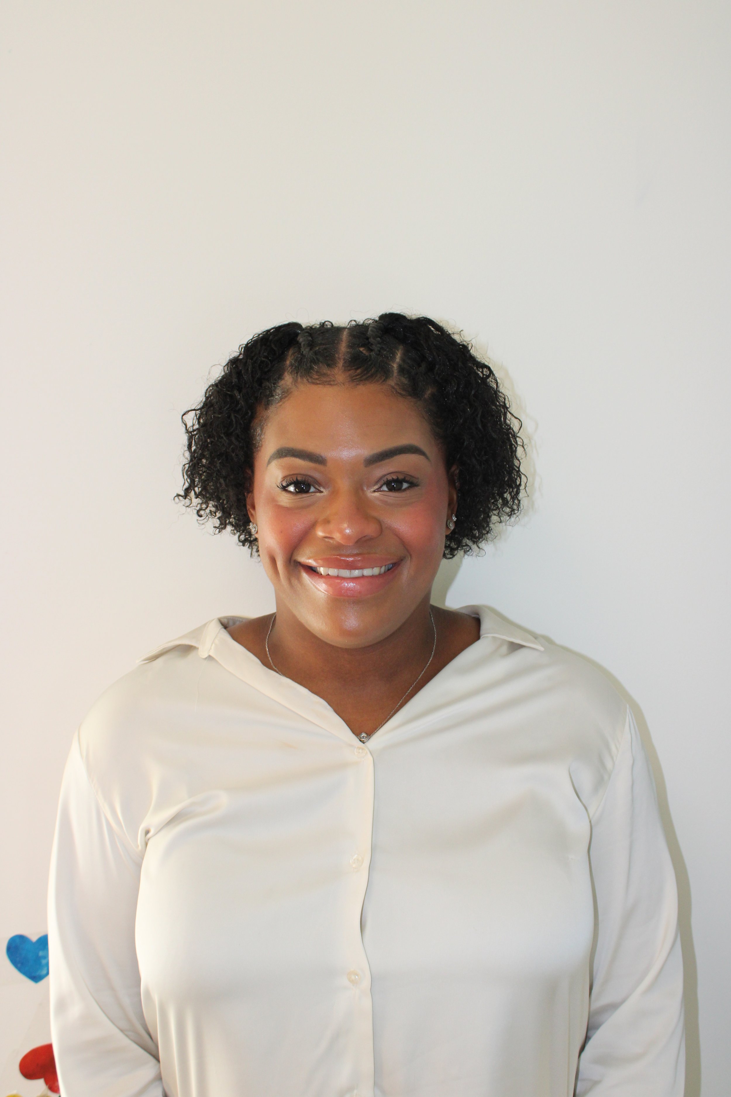 Smiling woman with black curly hair wearing a cream-colored satin blouse, standing against a plain white wall.