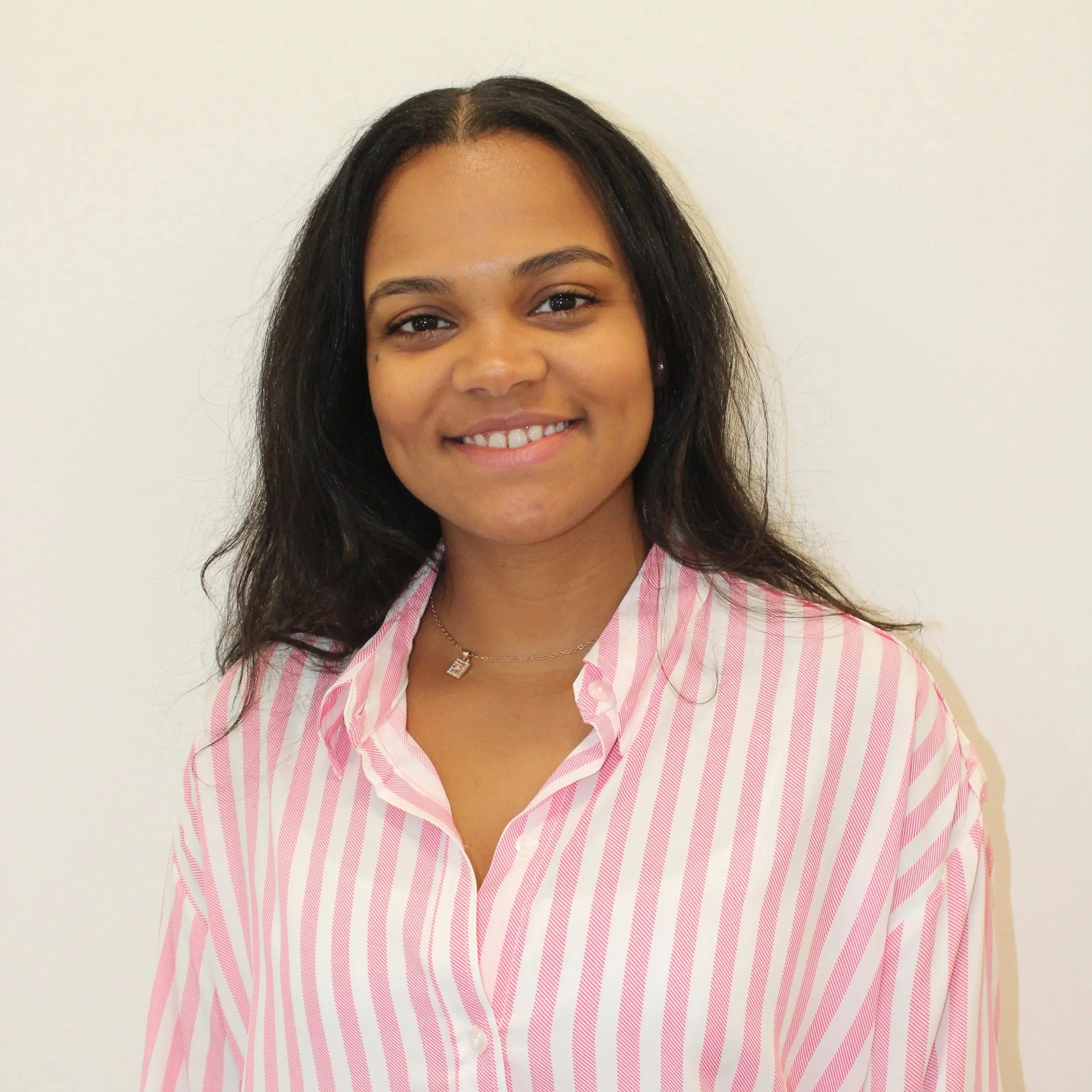 A woman with dark hair smiling, wearing a pink and white striped blouse and a necklace with a square pendant, standing against a plain light background.