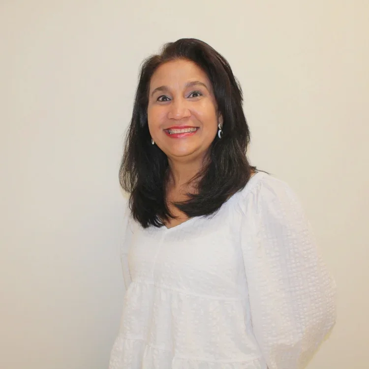 A smiling woman with dark hair wearing a white top, standing against a plain light-colored background.