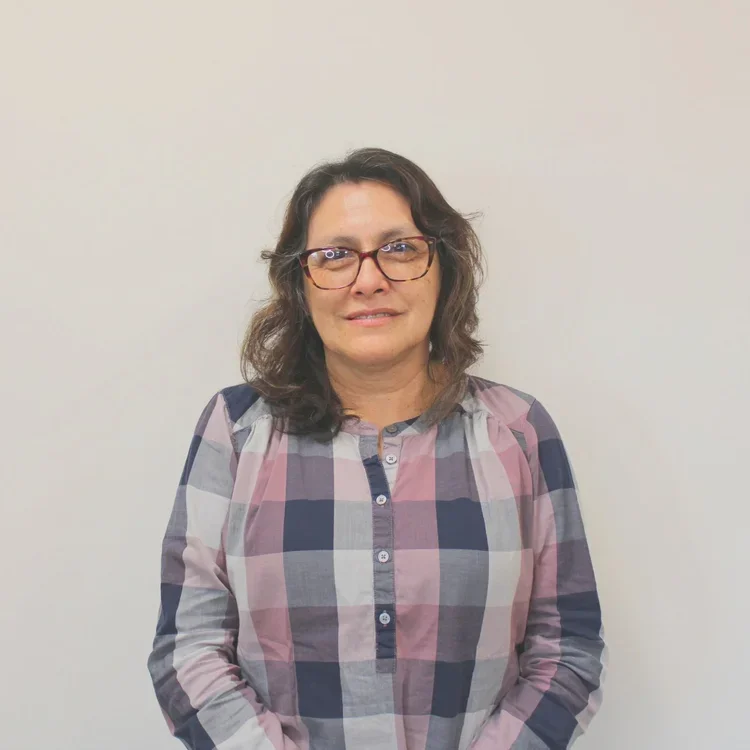 A woman with shoulder-length dark hair, wearing glasses and a plaid shirt, standing against a plain light-colored wall.