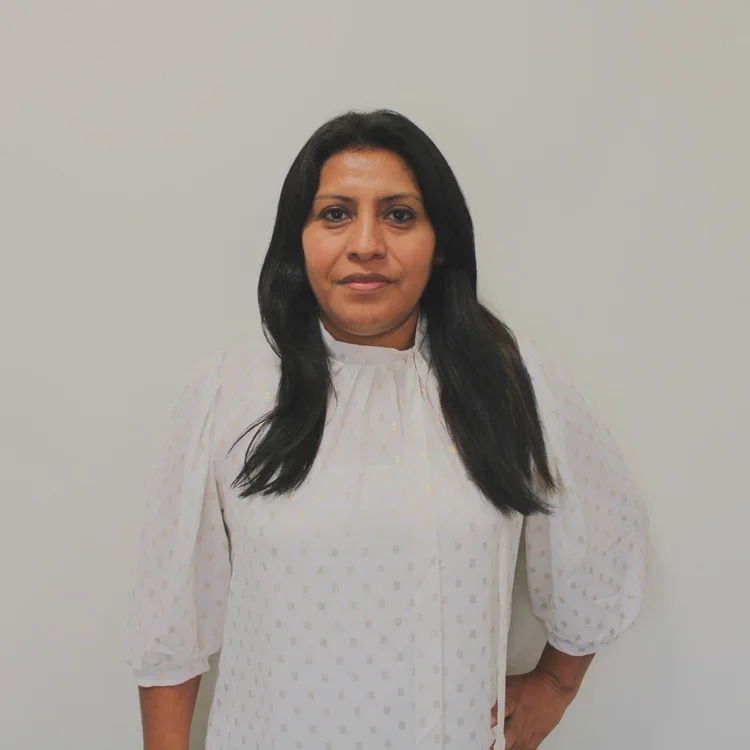 A woman with long black hair wearing a white dotted blouse standing against a plain gray wall.