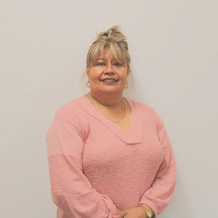 A woman with light blonde hair styled in an updo, wearing a pink long-sleeve sweater, gold jewelry, and standing against a plain white background.