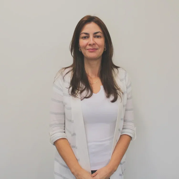 A woman standing against a plain white wall, wearing a white top and a light-colored cardigan, with long dark hair and a slight smile.