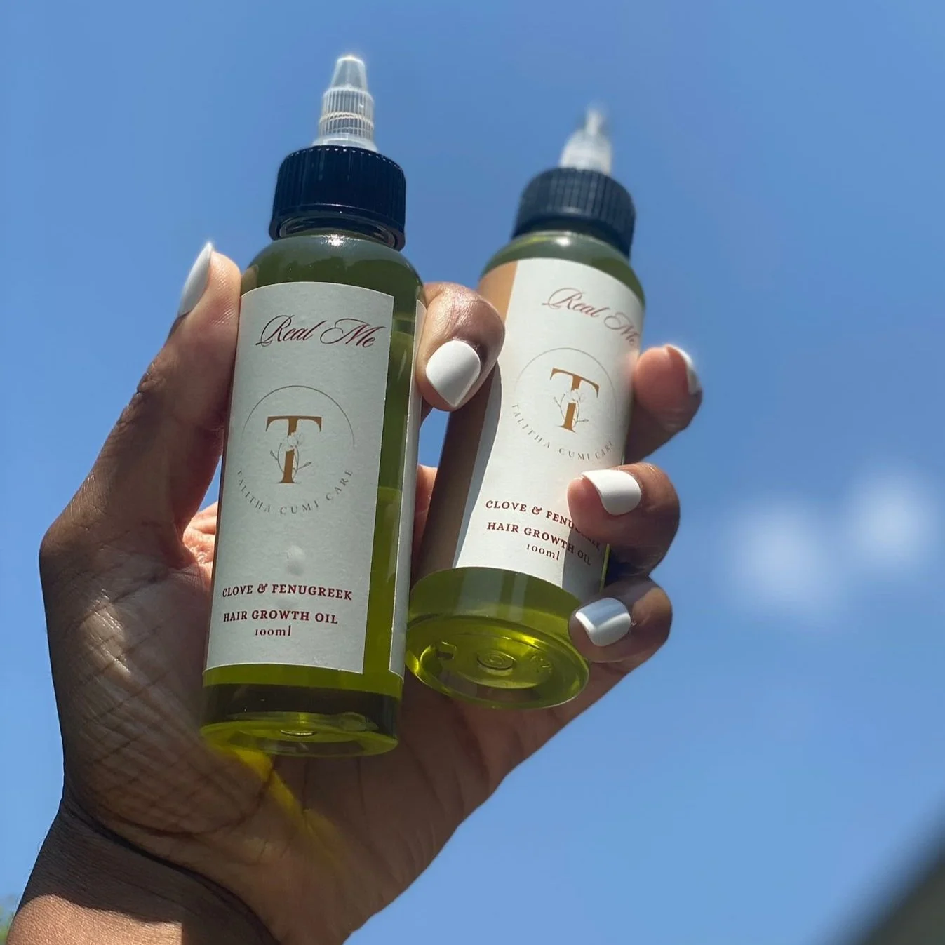 Two hands holding bottles of hair growth oil with blue sky in the background.