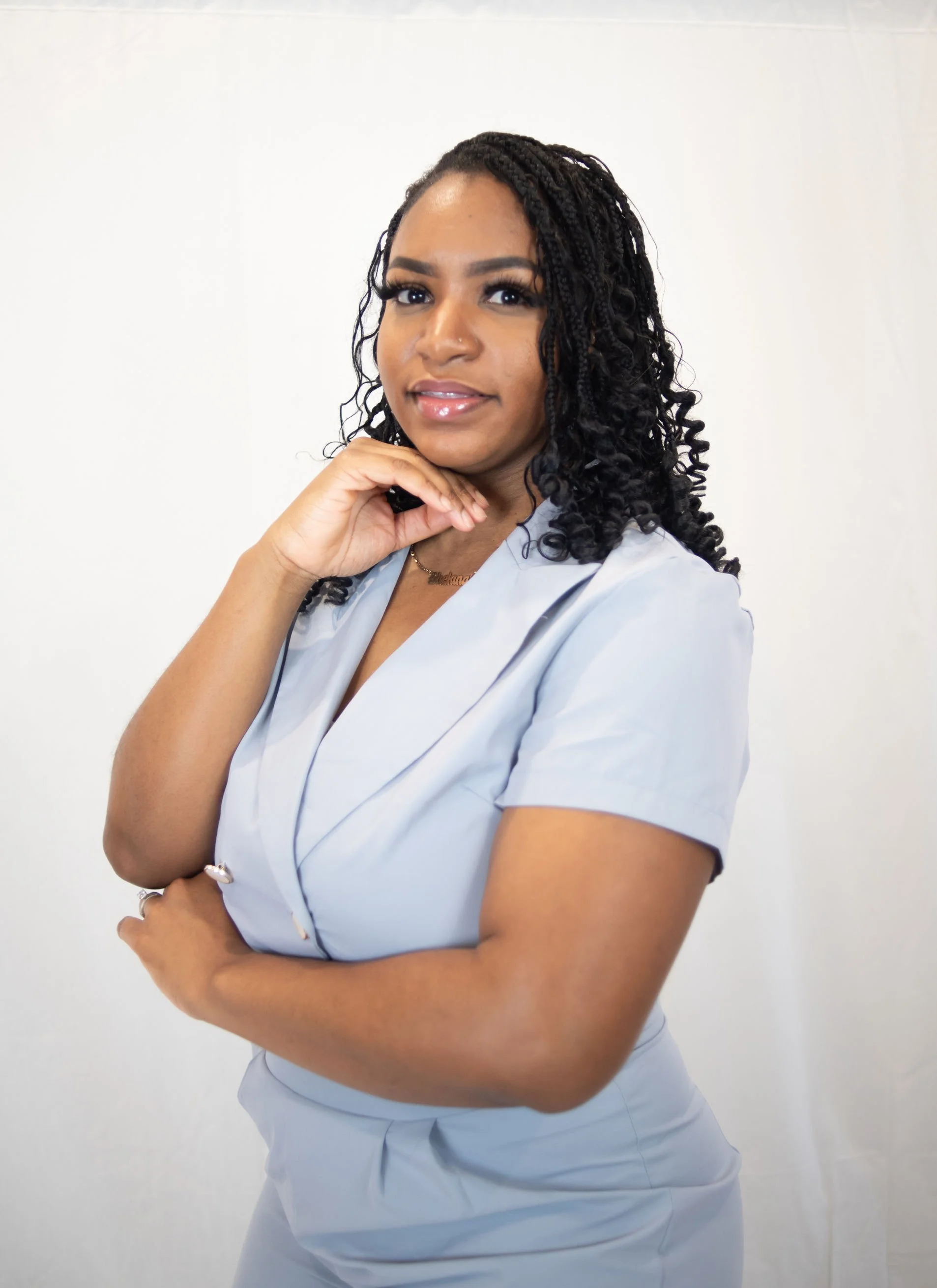 A young woman with curly hair wearing a light gray nursing uniform, standing against a plain white background, looking confidently at the camera with her hand resting near her chin.
