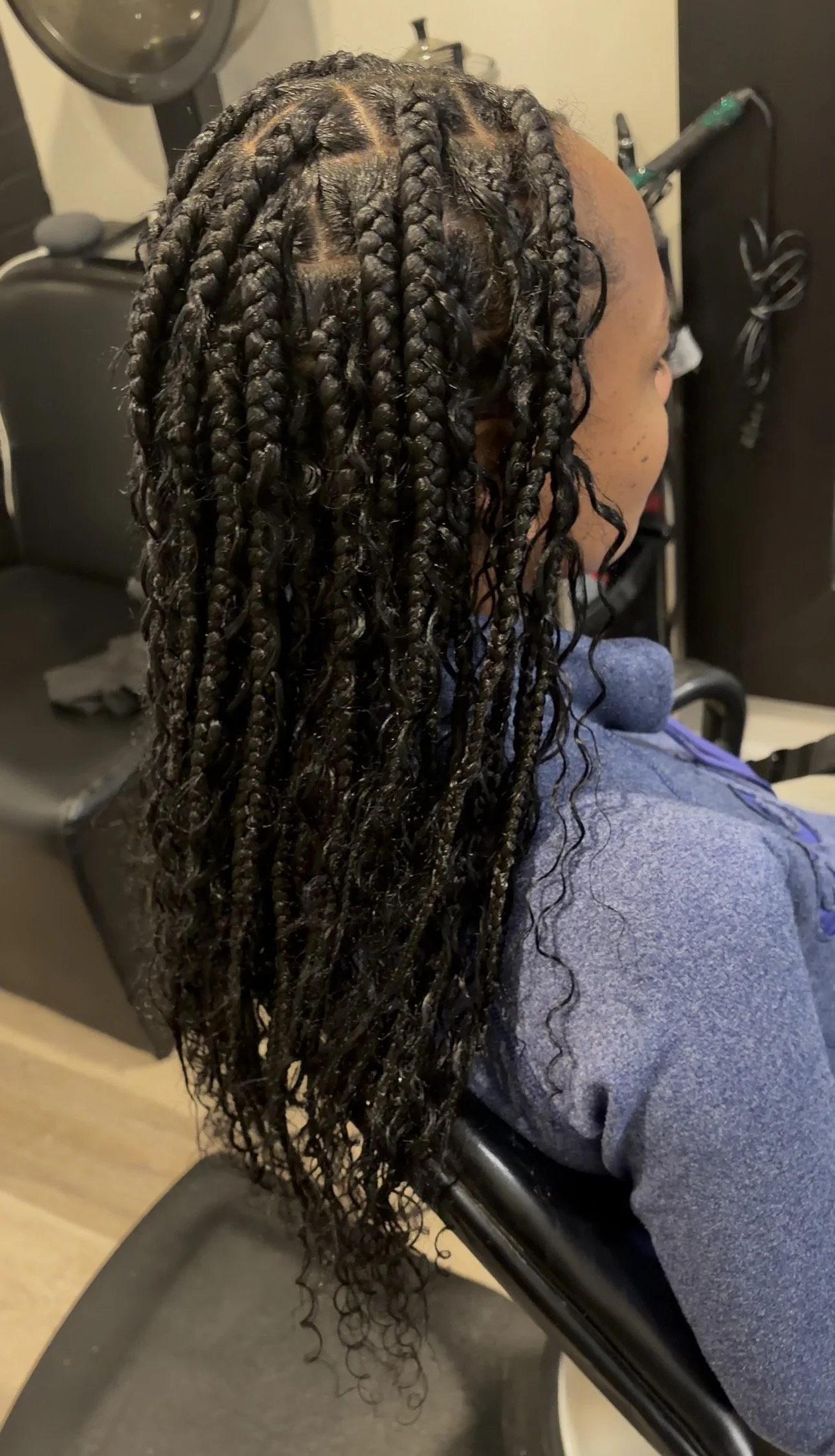Person with long, twisted braids in their hair, seen from the top, holding their hair back with both hands. The background is green foliage.