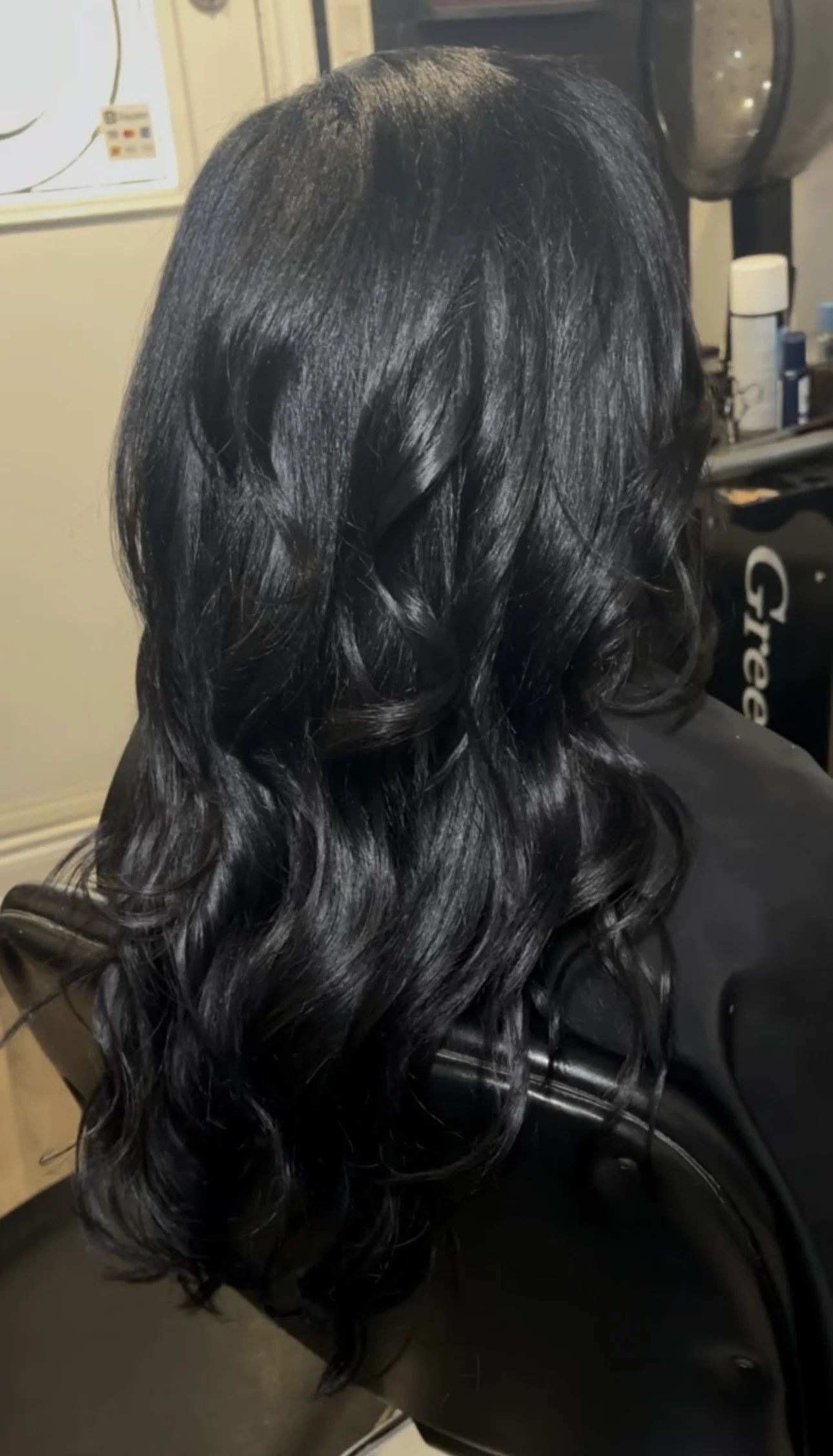 Close-up of a person's shiny, straight, black bob hairstyle with a slight angle, wearing a light pink Reebok shirt, against a green leafy background.