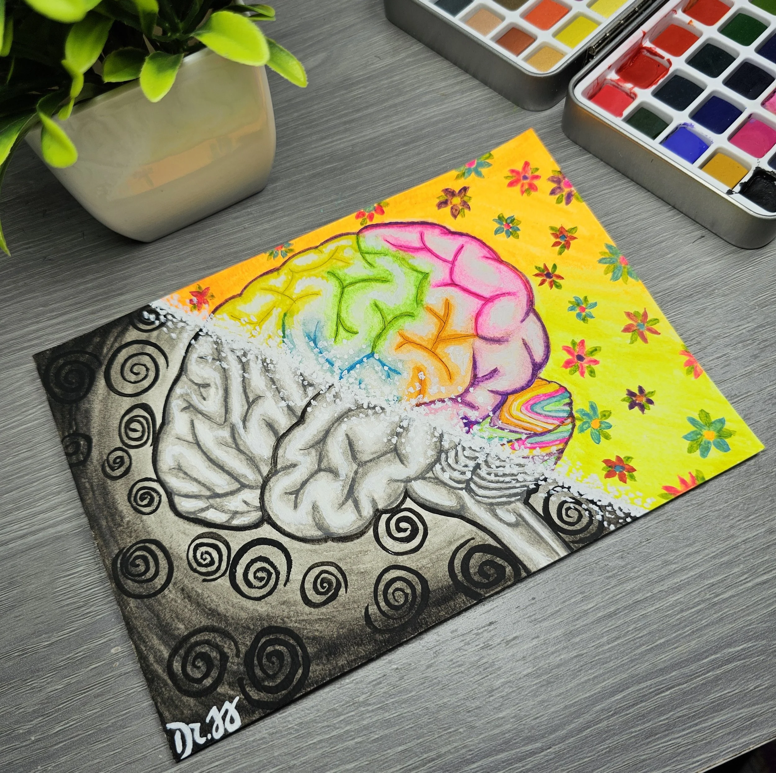 Colorful brain illustration on a yellow background, surrounded by small flower patterns, with watercolor paint sets and a plant visible on a wooden surface.