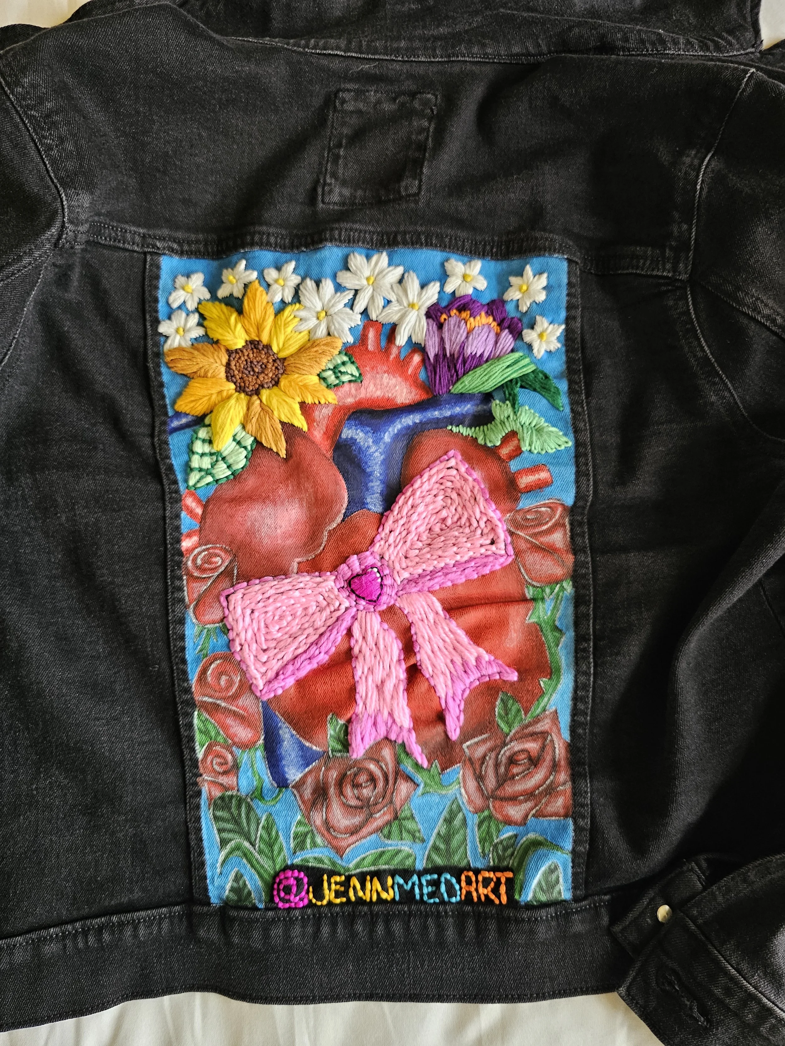 Black denim jacket with a colorful handmade embroidered patch on the back featuring flowers, a pink bow, and the Instagram handle @JENNMEDART in rainbow-colored text.