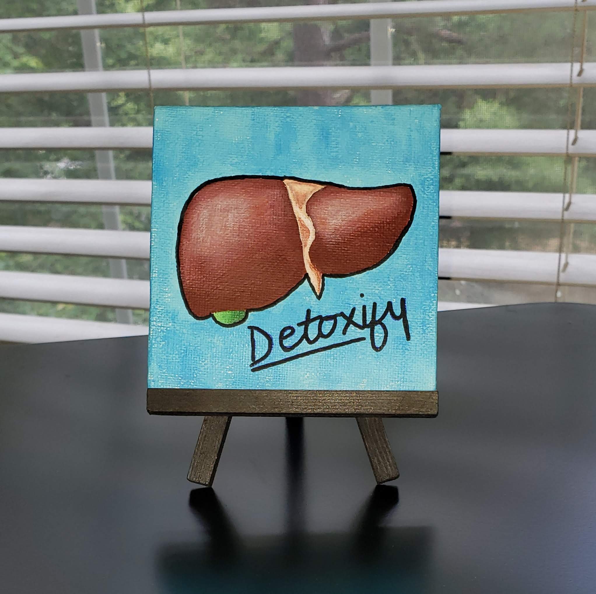 A small painting on a wooden easel featuring a brown liver. The background is painted in light blue with text below that spells 'detoxify'.