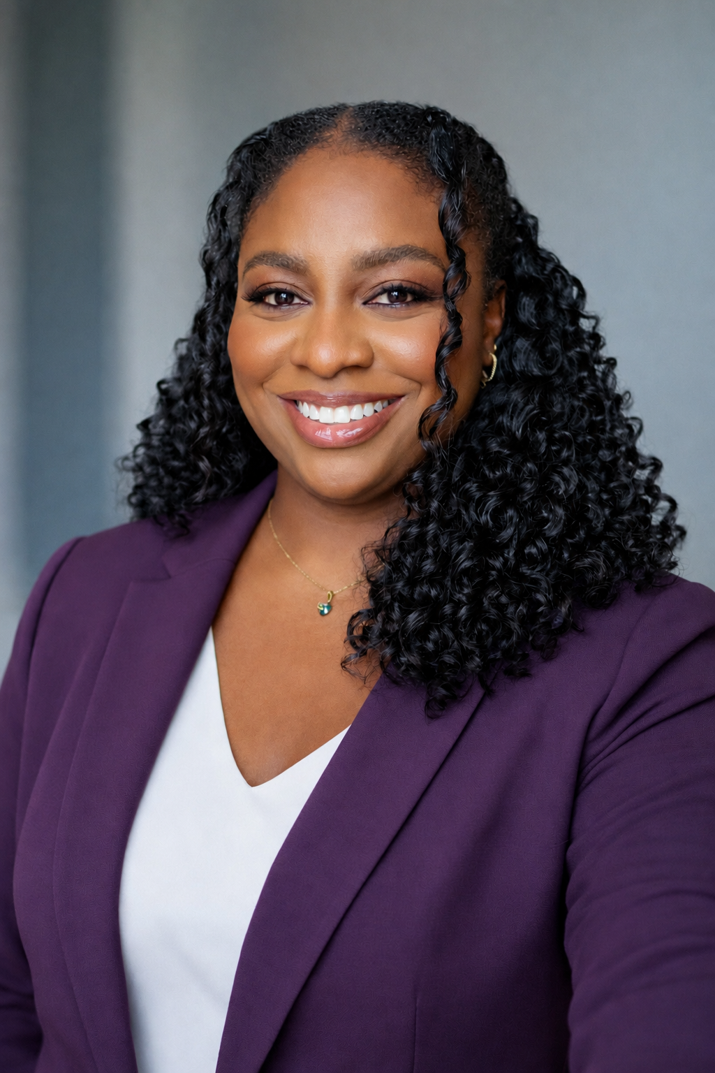 A professional woman with curly black hair, wearing a purple blazer, white top, and gold jewelry, smiling at the camera against a neutral background.