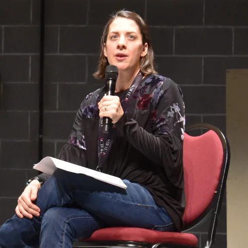 Image description: Talleri A. McRae sits on stage in a read chair talking to an audience holding a hand held microphone up to her mouth. Behind her is a concrete brick wall. She is wearing blue jeans and a black blouse with an embroidered floral print.