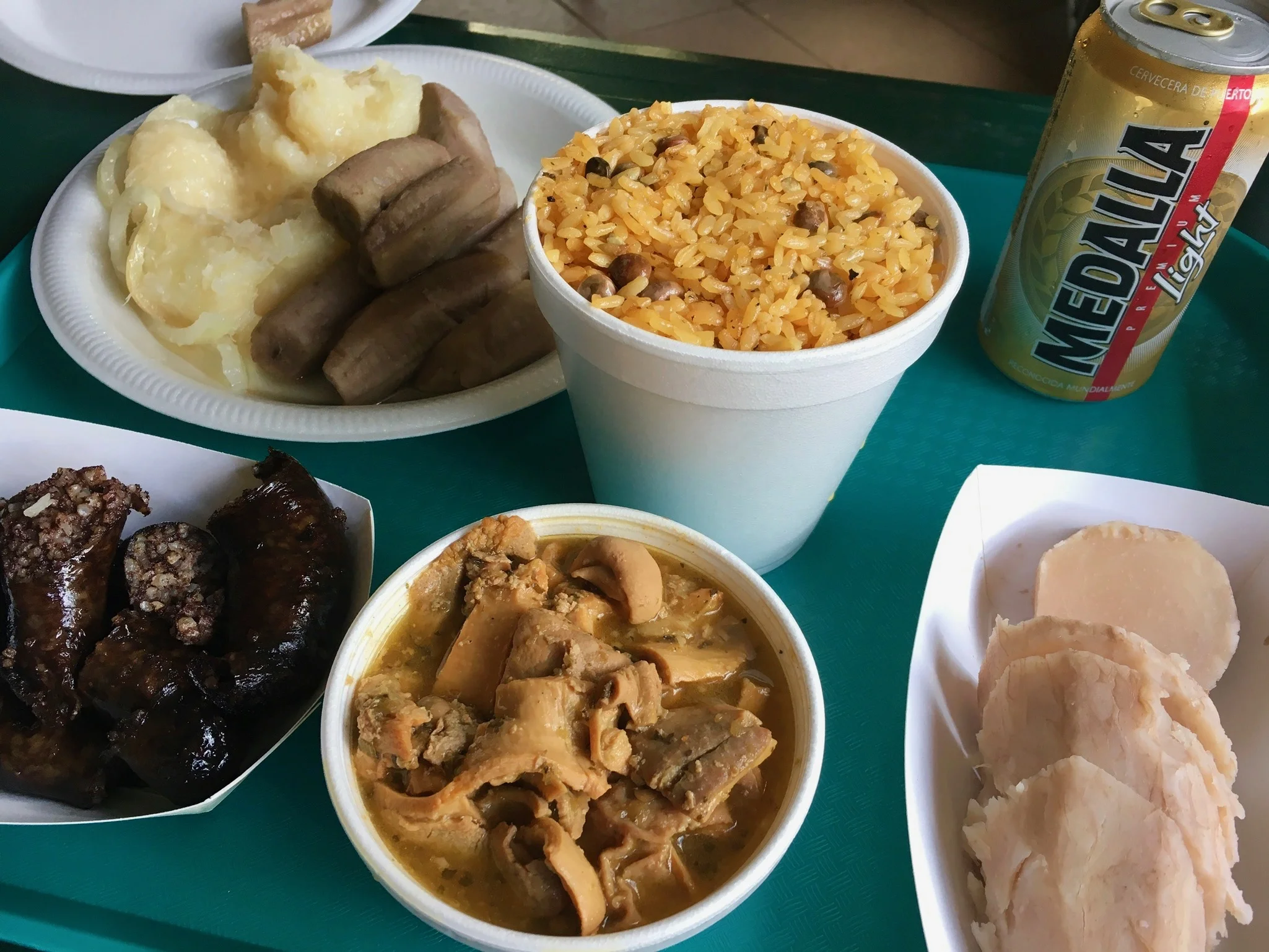 A typical spread of fixings from a lechonera. From left to right: stewed yuca, boiled guineo (green banana), arroz con gandules, a cold Medalla, yautia (taro root), cuajito (stewed pig stomach), and morcilla (fried blood sausage).