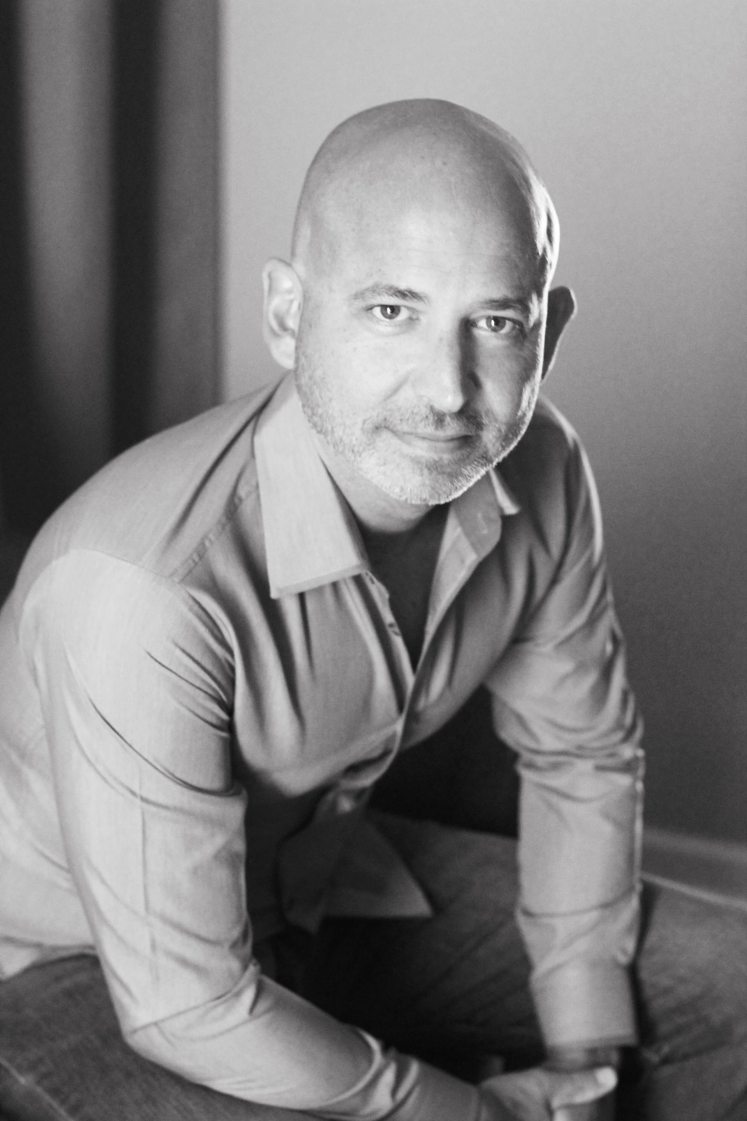 A black-and-white photo of a man with a shaved head and beard, dressed in a collared shirt, sitting on the floor with his right arm resting on his knee, looking at the camera.