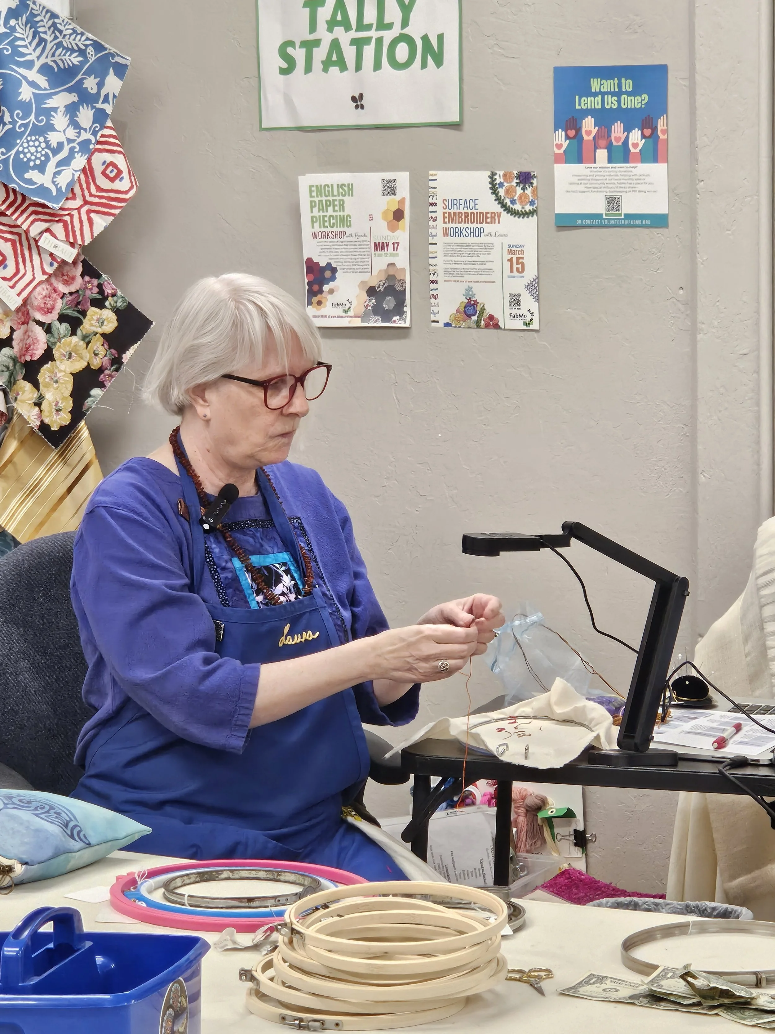 Seated woman holding needle and fabric under small camera in embroidery demonstration