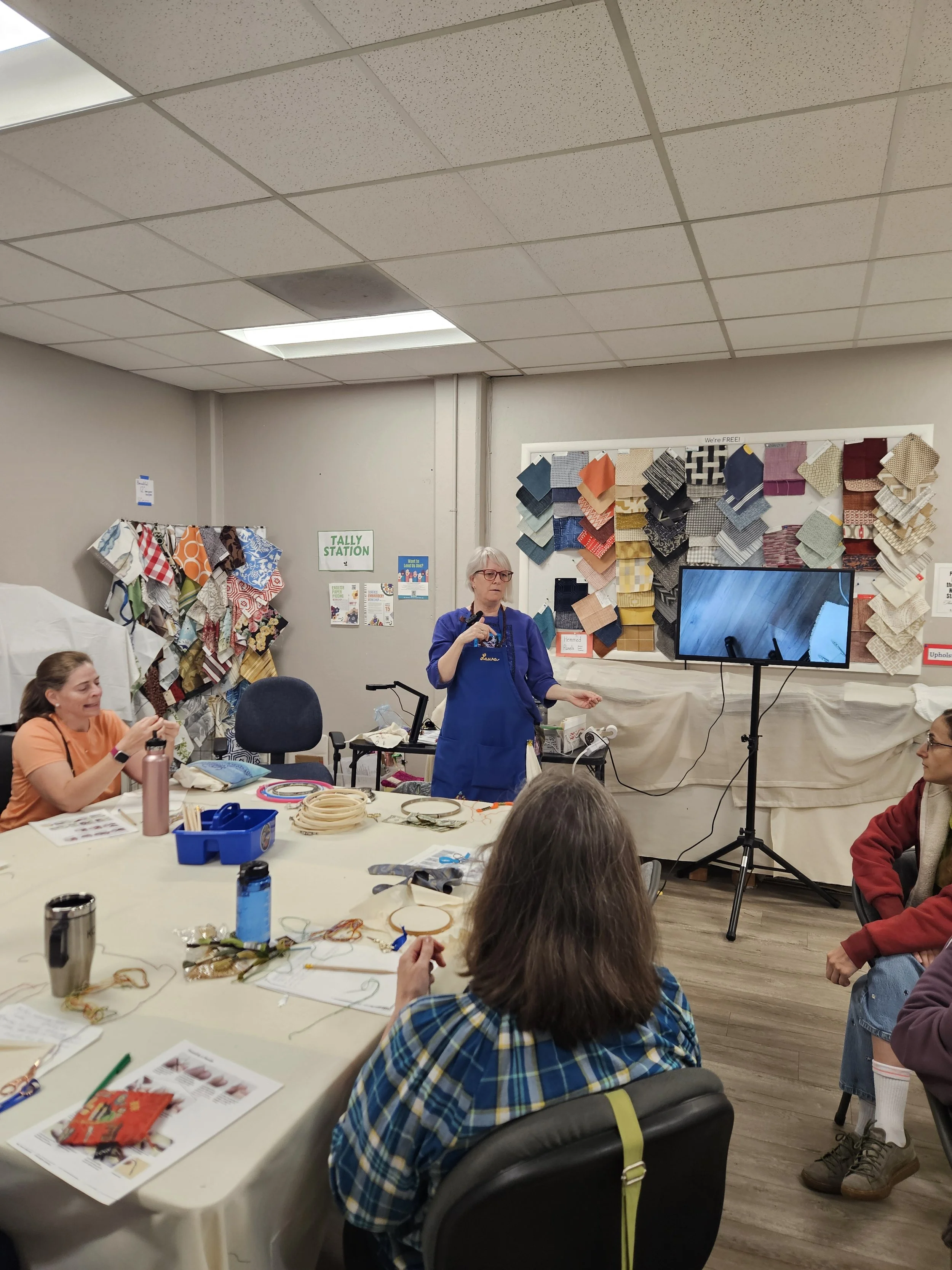 Woman standing in front of table of seated people, next to monitor showing embroidered fabric