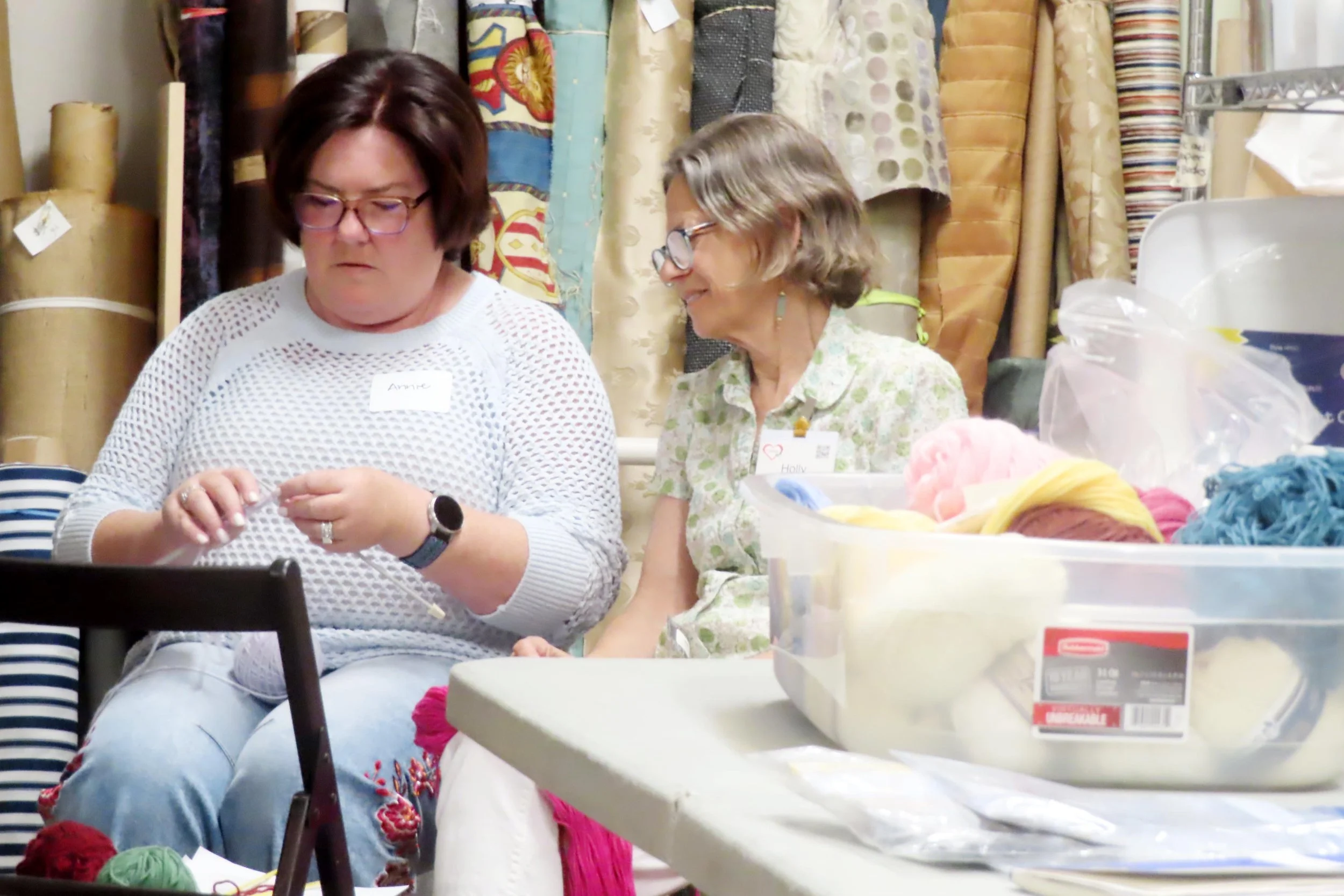 Two women seated side by side, one looking at the other woman holding knitting needles