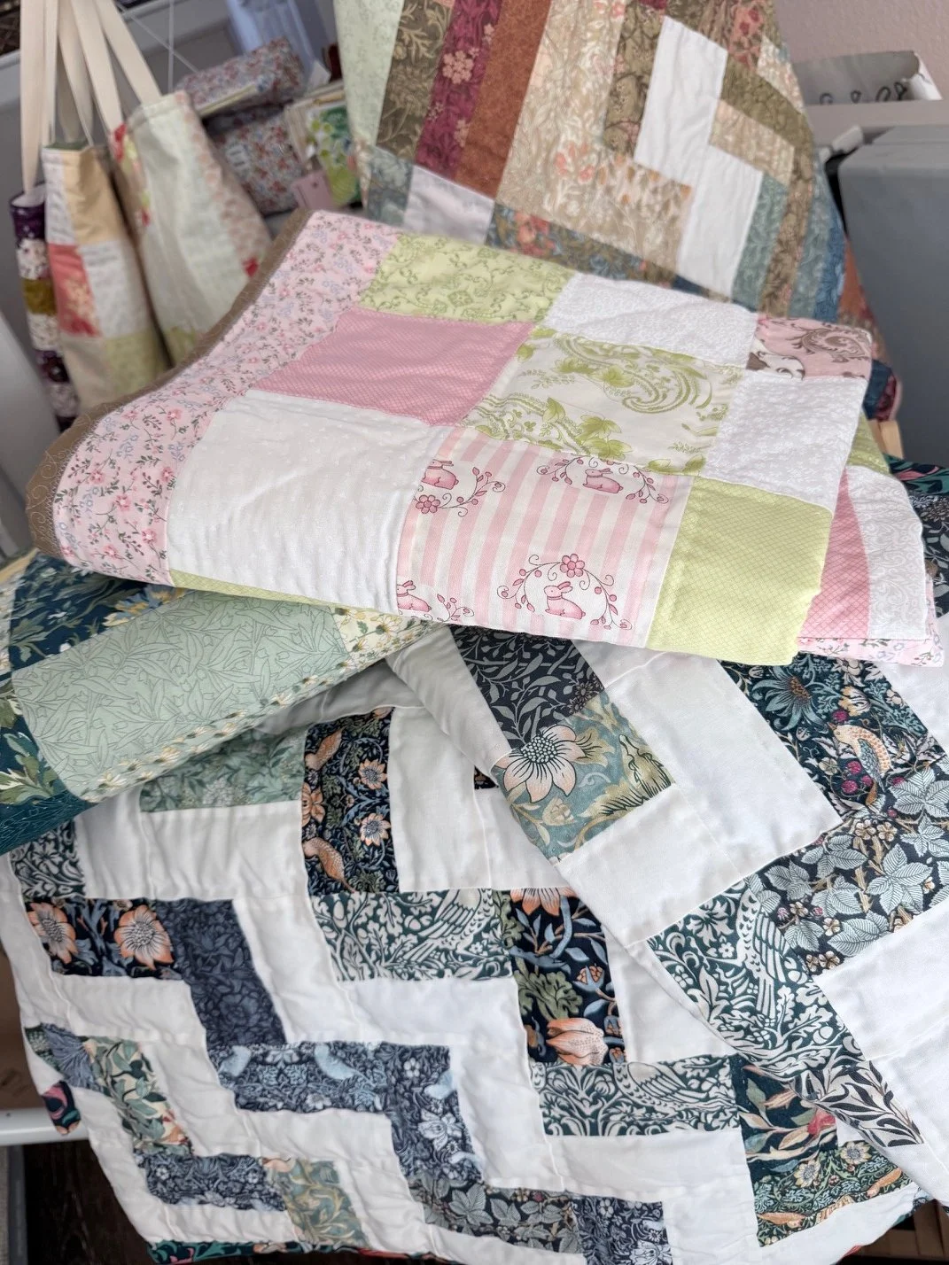 Pile of handmade quilts with patchwork tote bags in the background