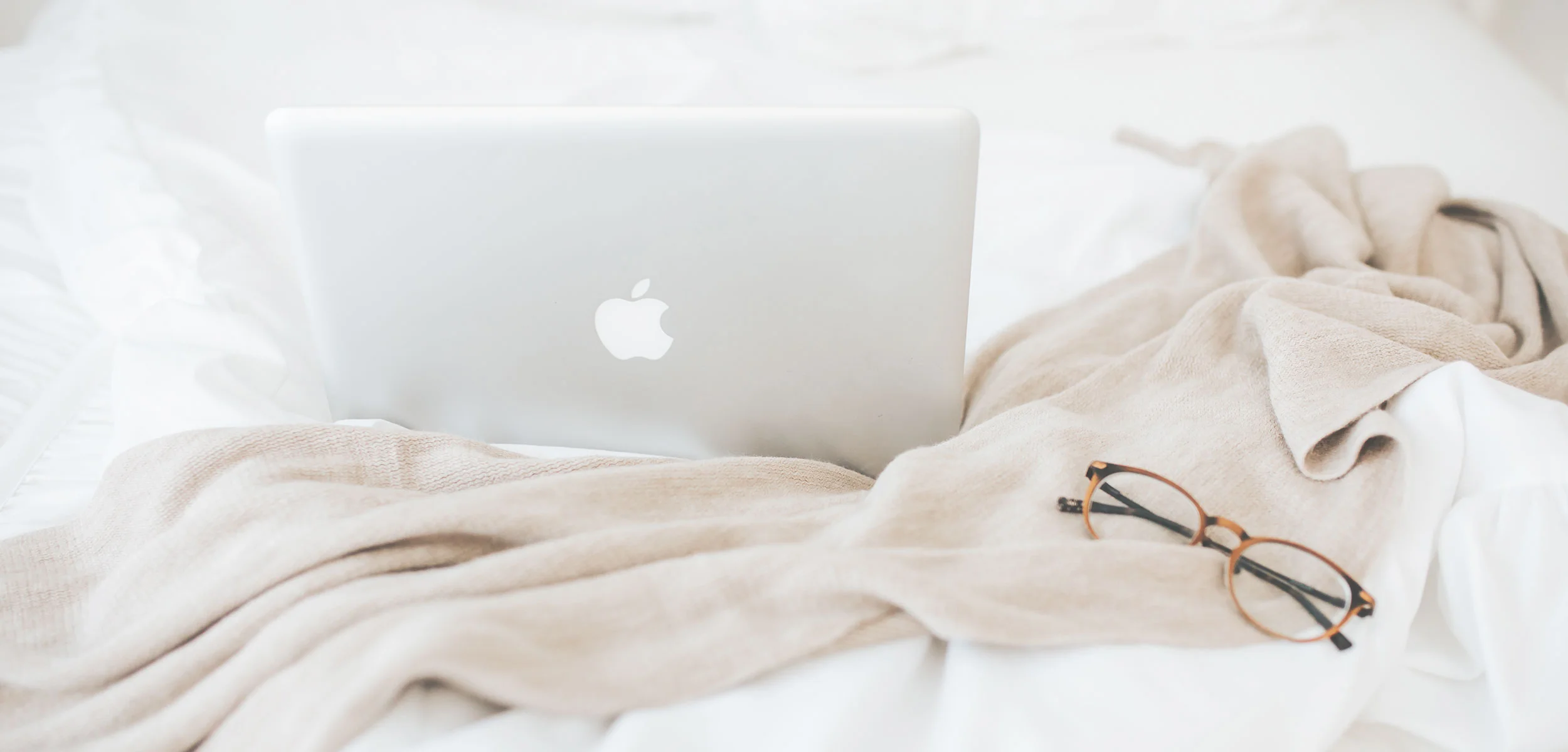 Macbook sitting on top of a tan blanket on a white bed, with glasses sitting beside it
