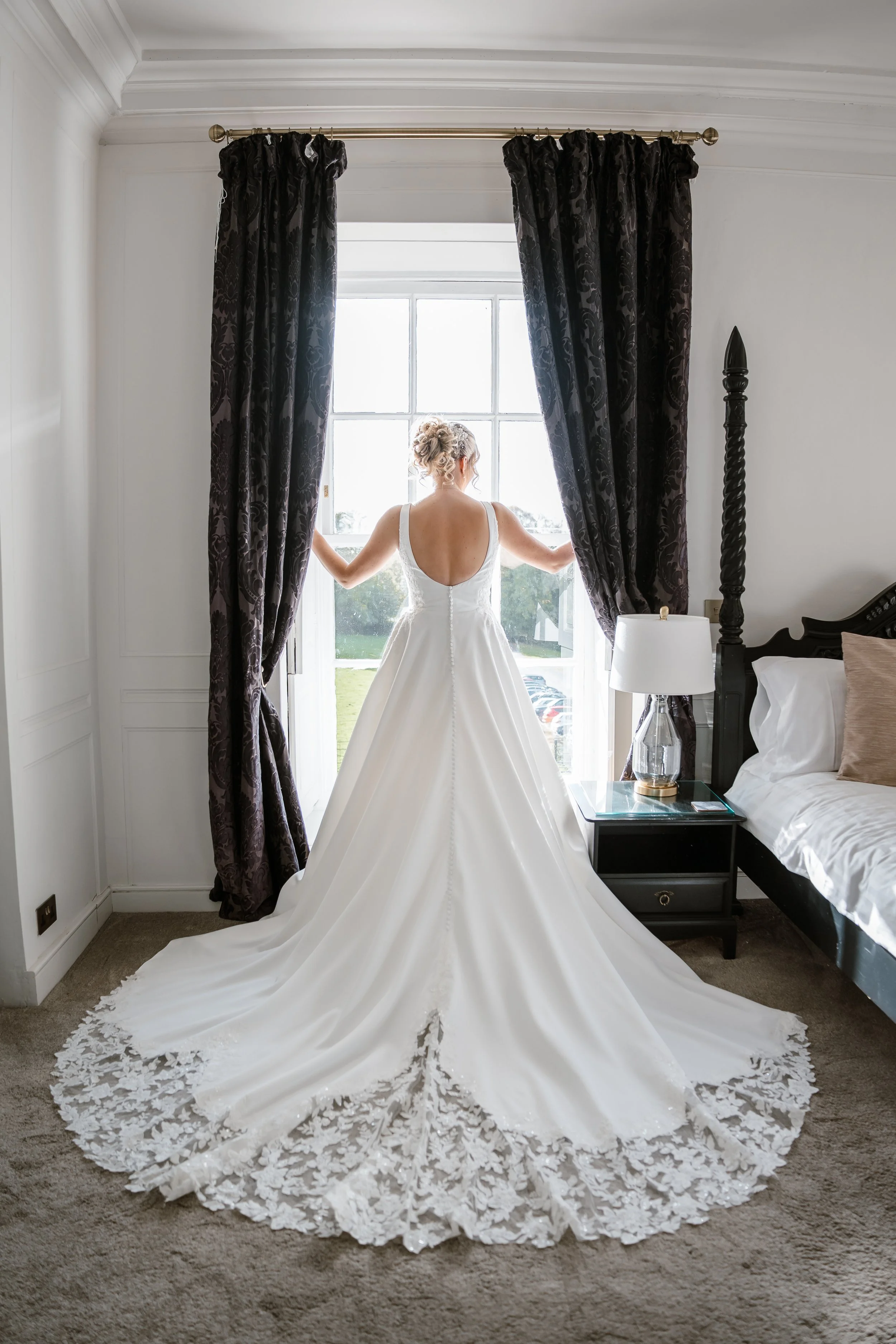 A bride in a white wedding gown stands in front of a large window, holding the curtains open, with a view of a green outdoor area.