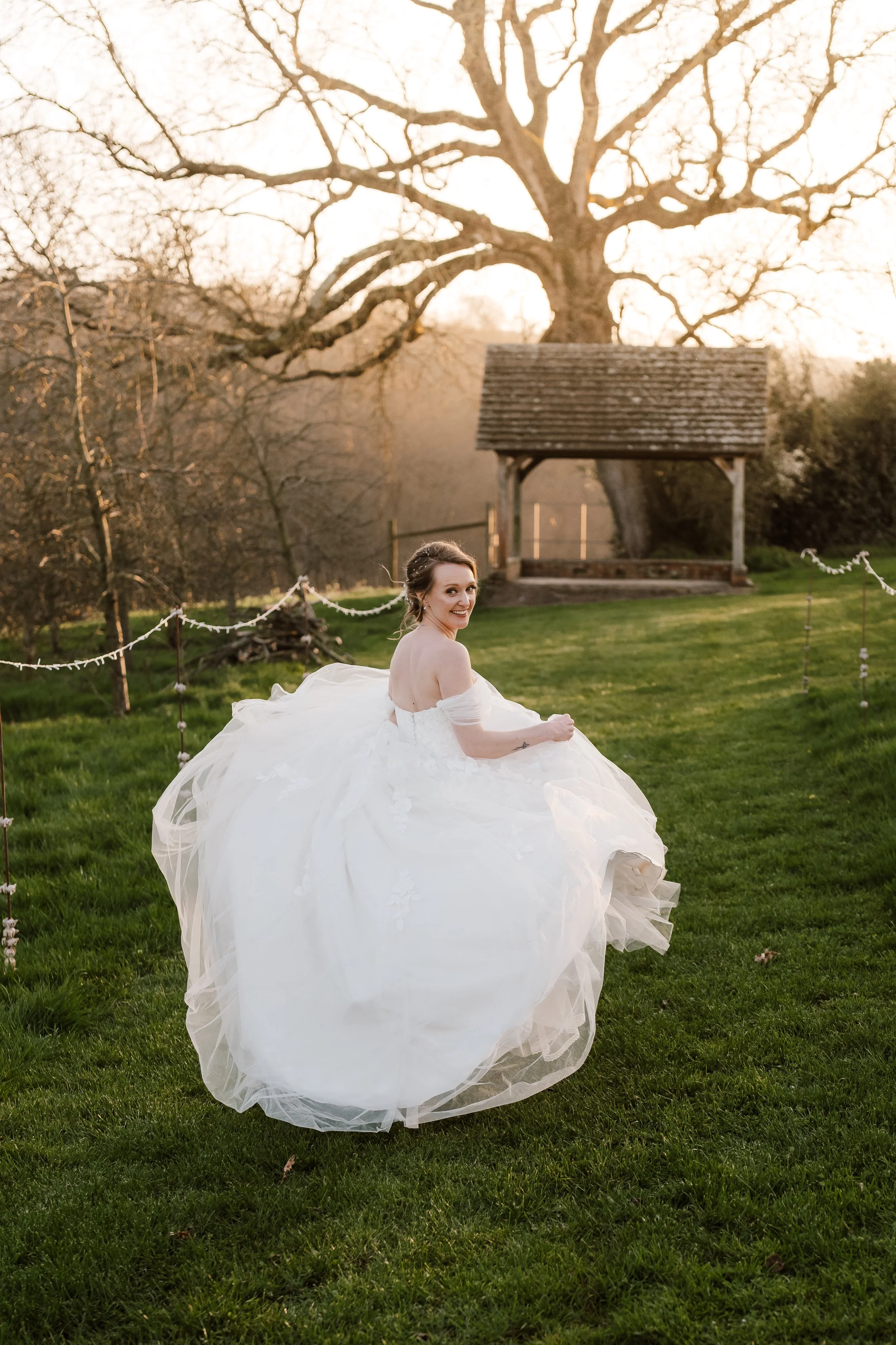A woman in a wedding dress sitting on grass in a garden with a large tree and a small wooden structure in the background, during sunset.