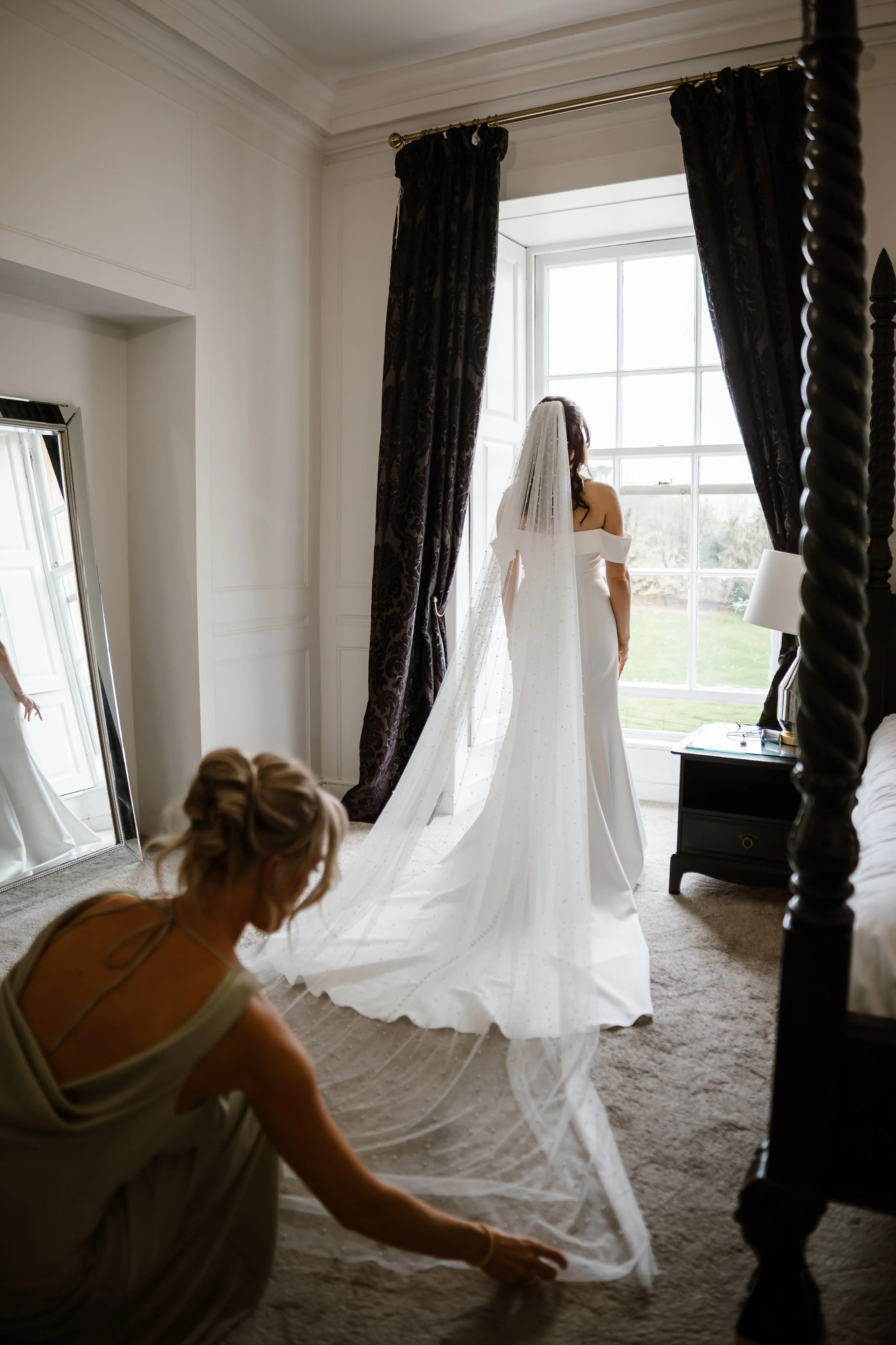 A bride standing in front of a large window with a view outside, with her back to the camera, while a woman adjusts her wedding veil on the floor in a well-lit bedroom.