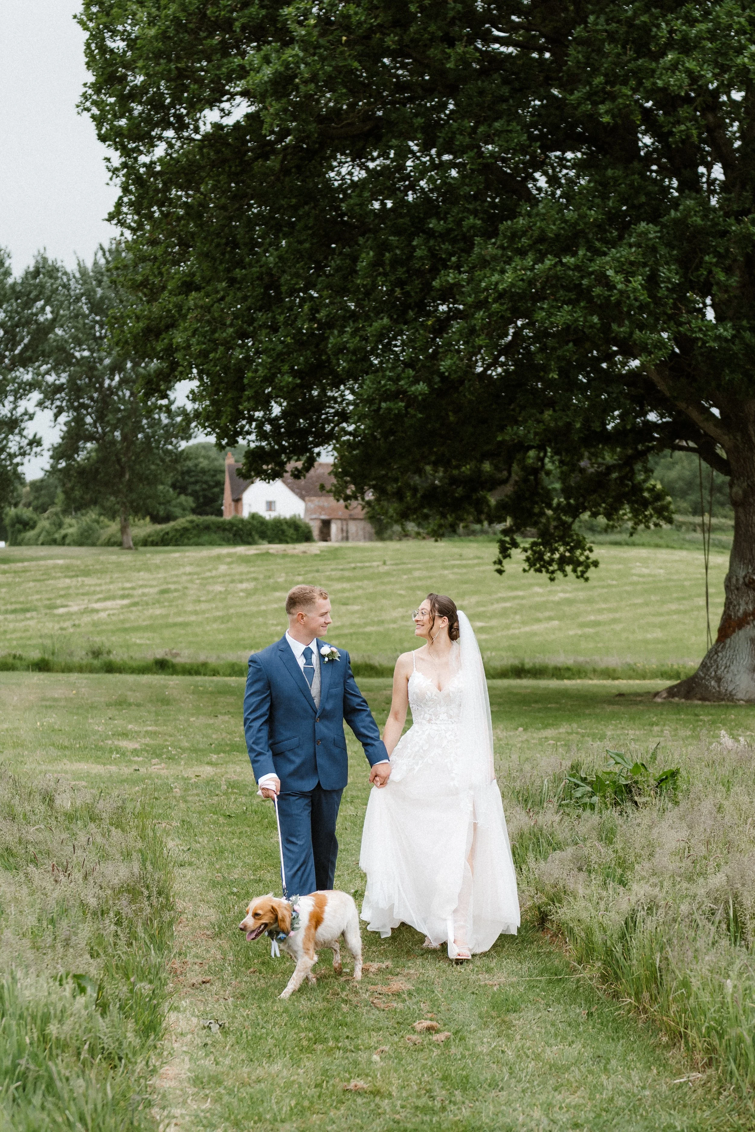 A bride and groom holding hands and walking outdoors on a grassy path, accompanied by a dog, under a large tree with a country house in the background.