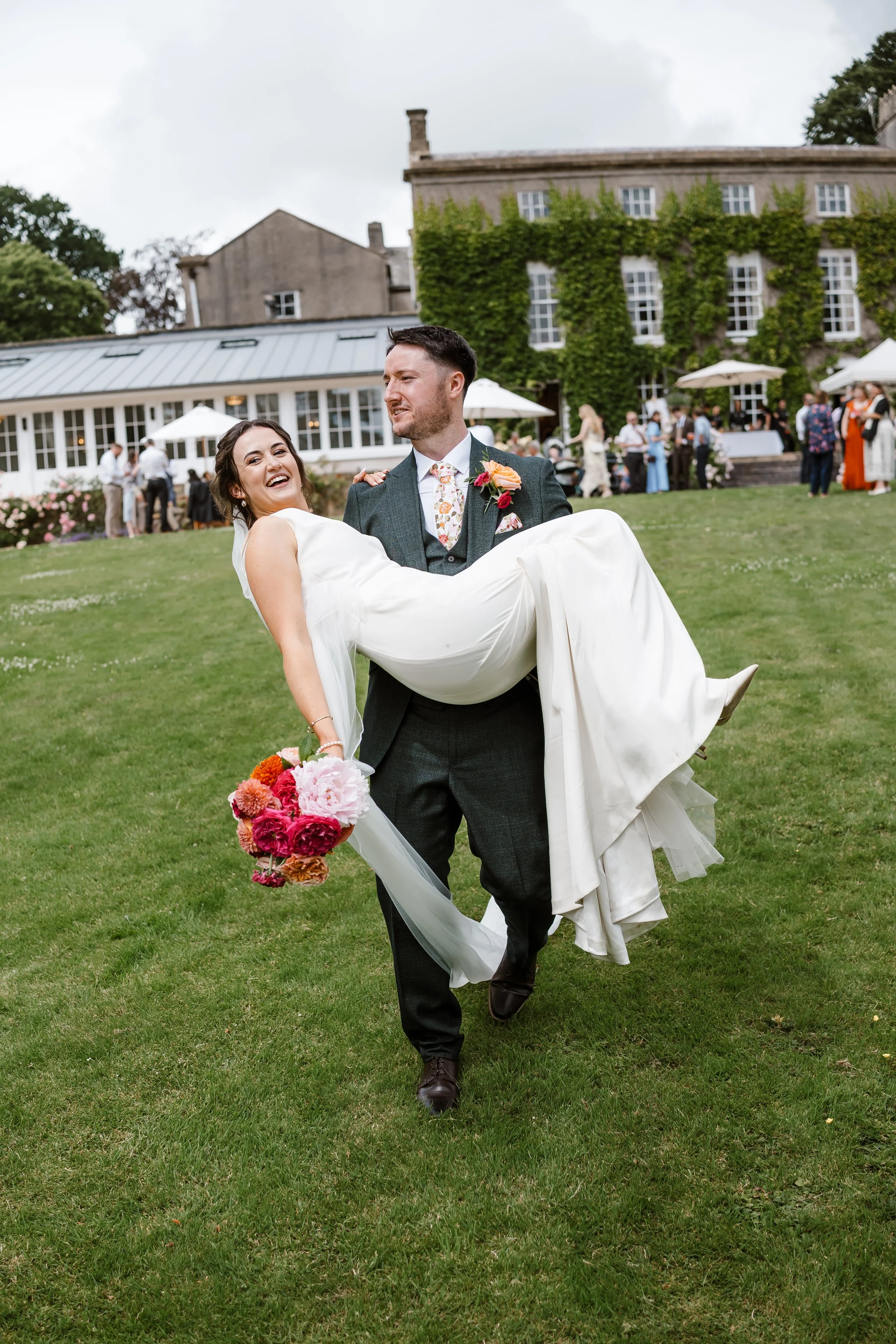 A wedding couple enjoying their outdoor wedding celebration on a grassy lawn outside a large building covered in green vines. The groom is holding the bride in his arms, and she is holding a bouquet of pink, red, and orange flowers.