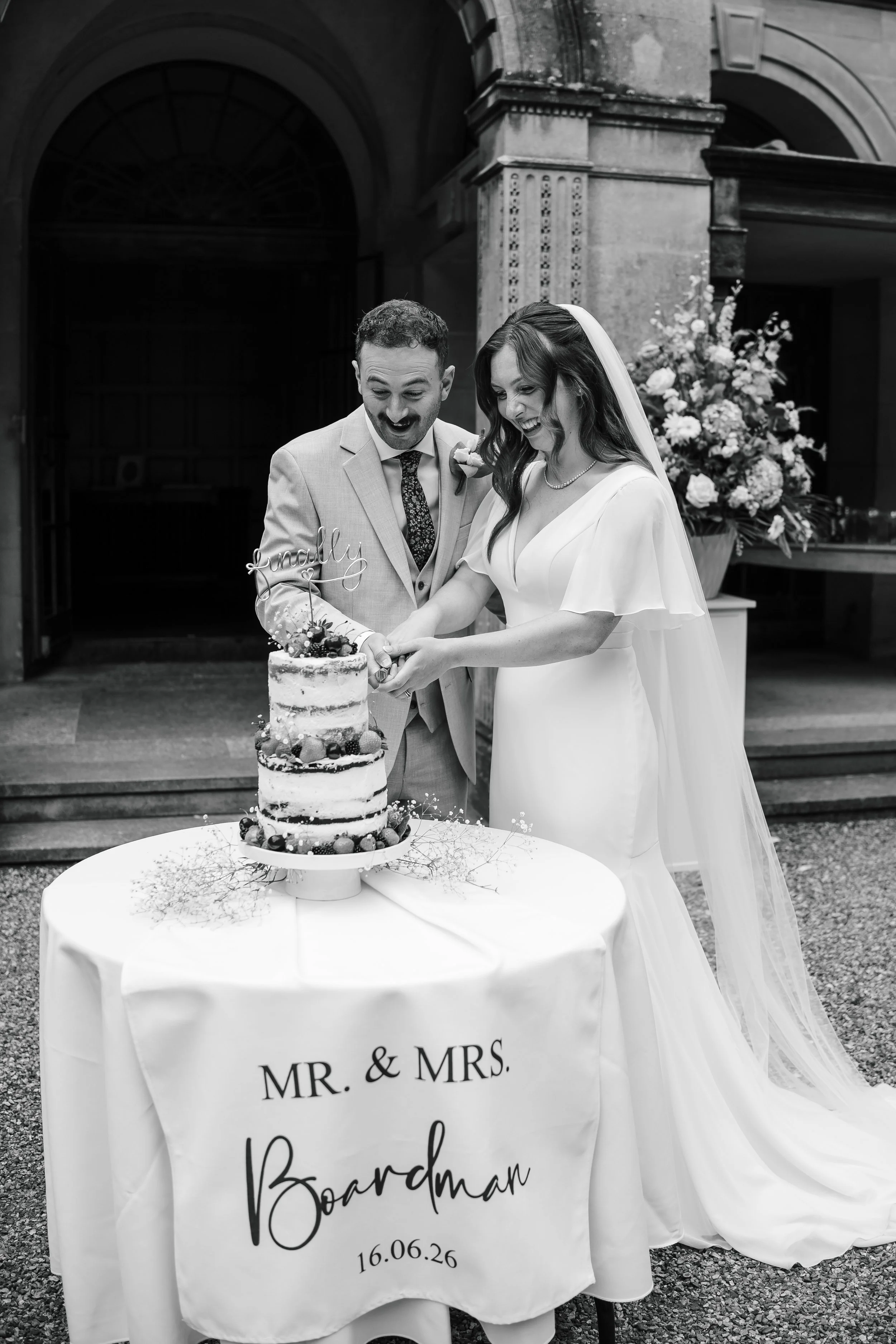 A wedding celebration with a bride and groom cutting a wedding cake in front of a decorated table with a 'Mr. & Mrs.' banner. The bride wears a white dress and veil, and the groom is in a light suit. A large floral arrangement is in the background.