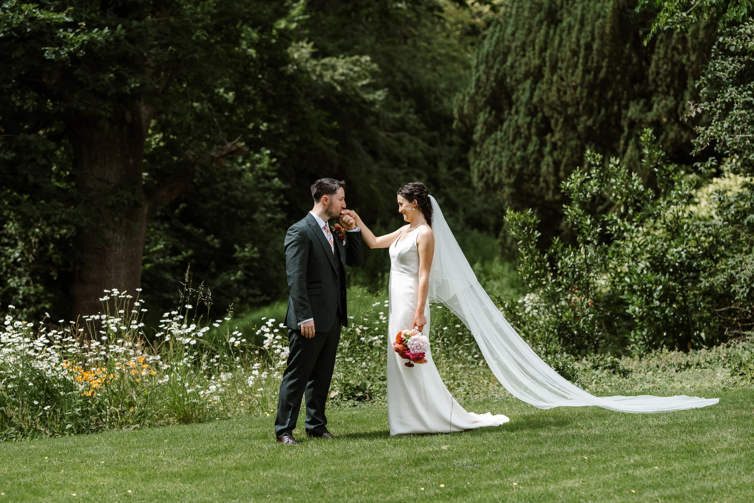 A bride and groom standing in a lush green outdoor setting with trees and flowers, sharing a moment during their wedding day.