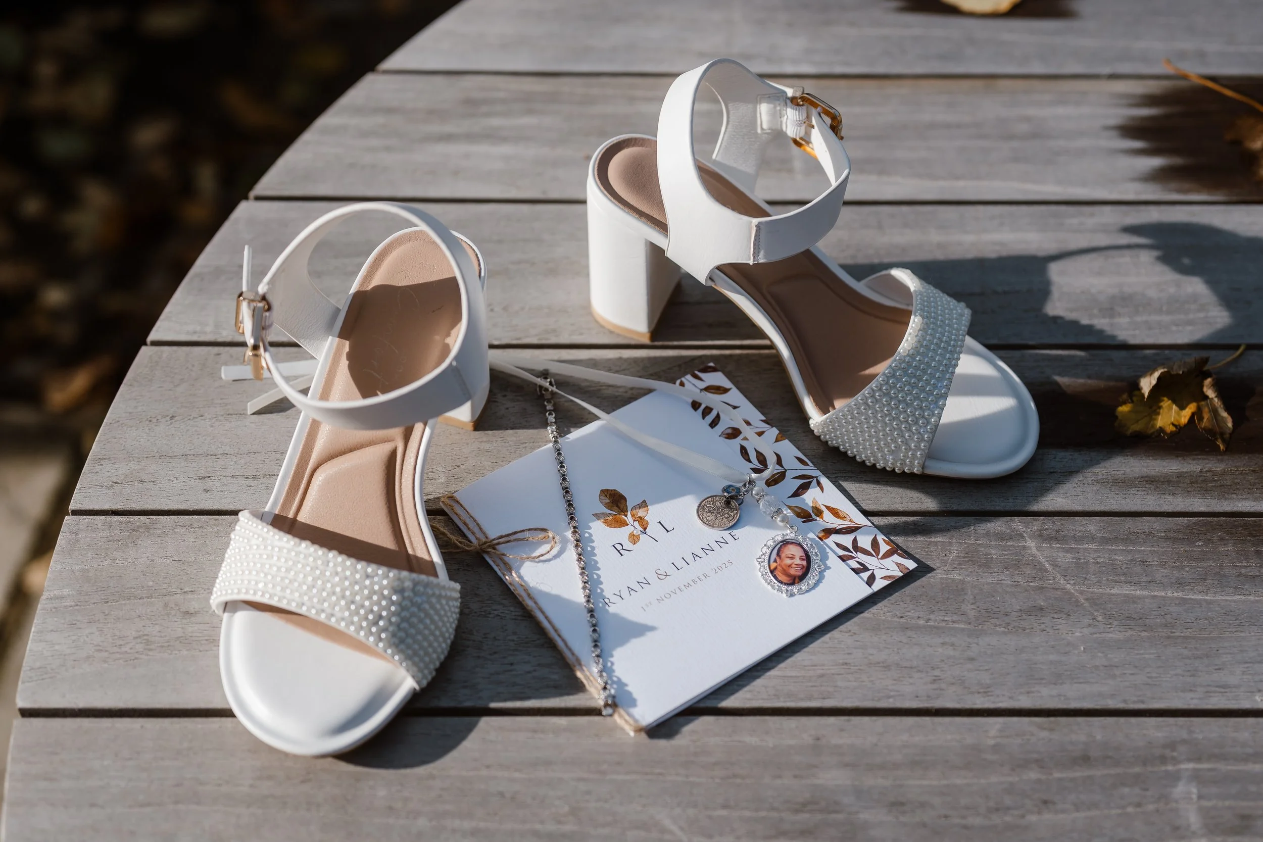 White heeled sandals with ankle straps and pearl embellishments, a wedding invitation with names Ryan and Lianne, and a silver necklace with a round photo pendant, all set on a wooden surface outside.