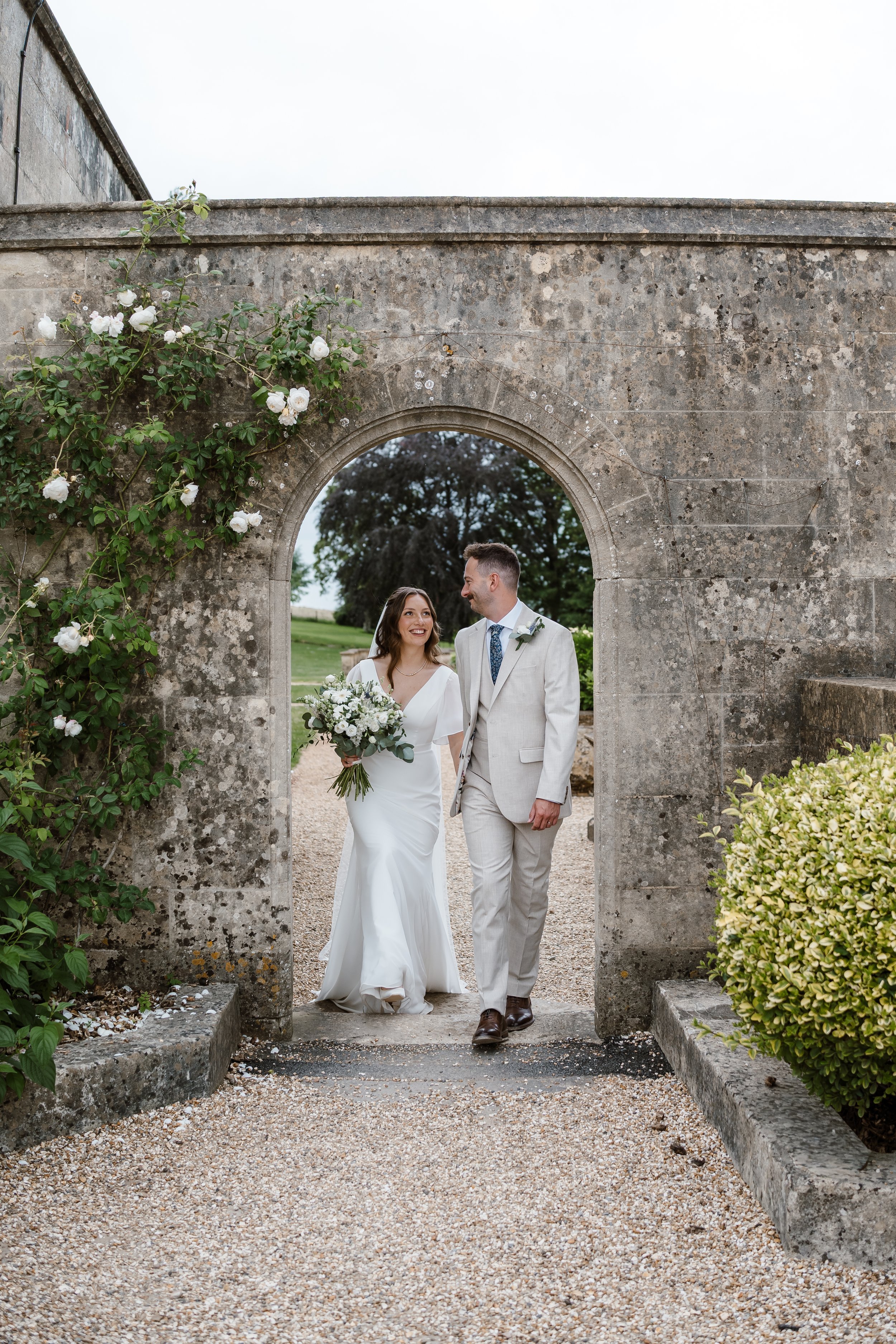 A bride and groom walk through a stone archway in an outdoor garden setting. The bride holds a bouquet of white and green flowers, and sisters in elegant wedding attire walk behind them, smiling and looking at each other.