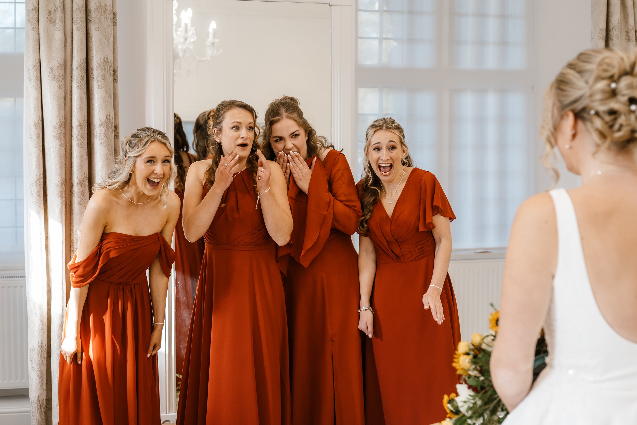 A bride in a white wedding dress holding a bouquet faces five bridesmaids in matching burnt orange dresses, all reacting with excitement and surprise.