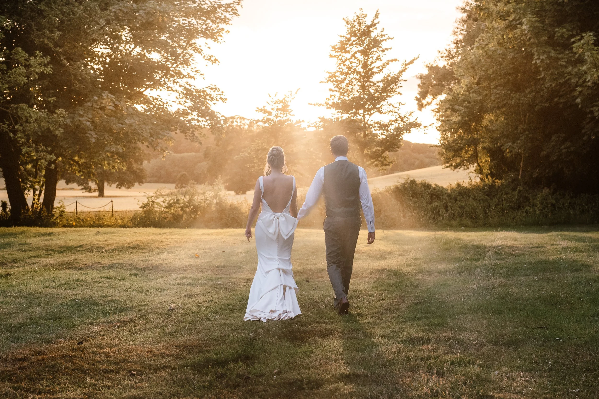 A bride and groom walking hand in hand in a grassy field during sunset, surrounded by trees.