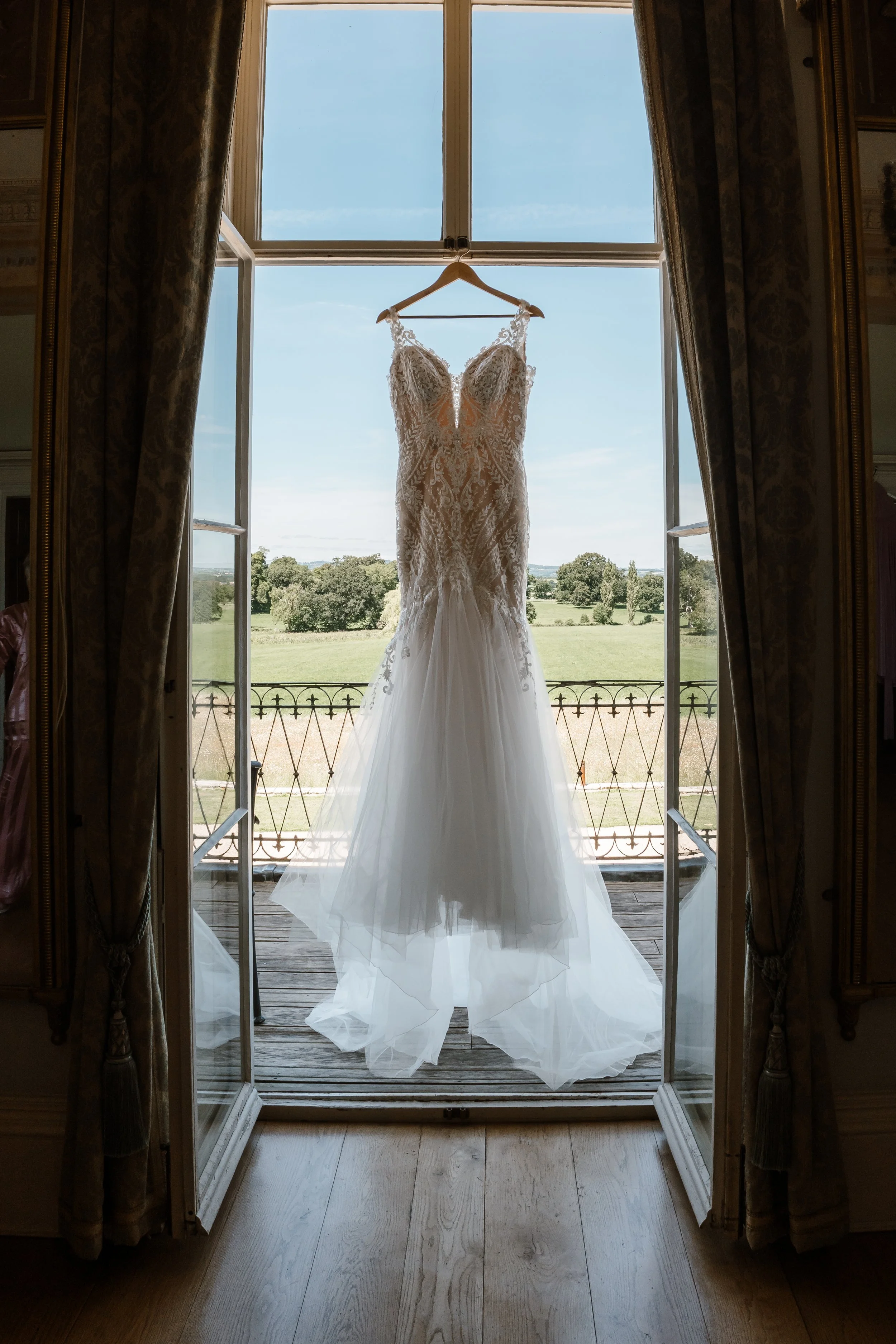 A white wedding dress hanging on a hanger in front of a large open window, with a scenic green landscape and blue sky outside.