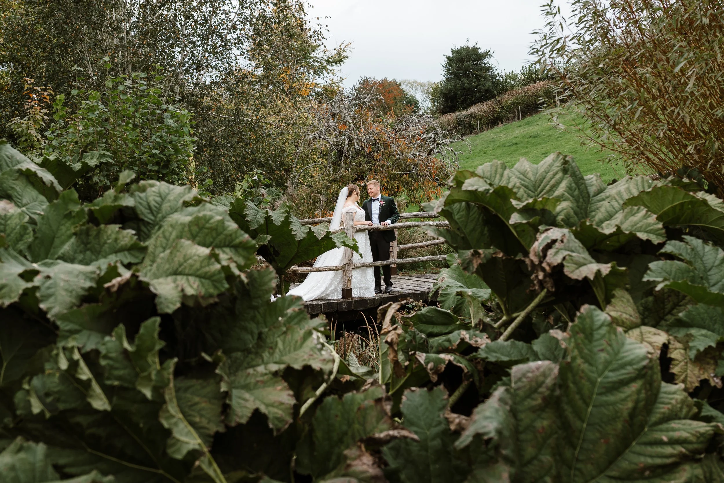 A bride and groom standing on a wooden bridge surrounded by lush greenery and trees.