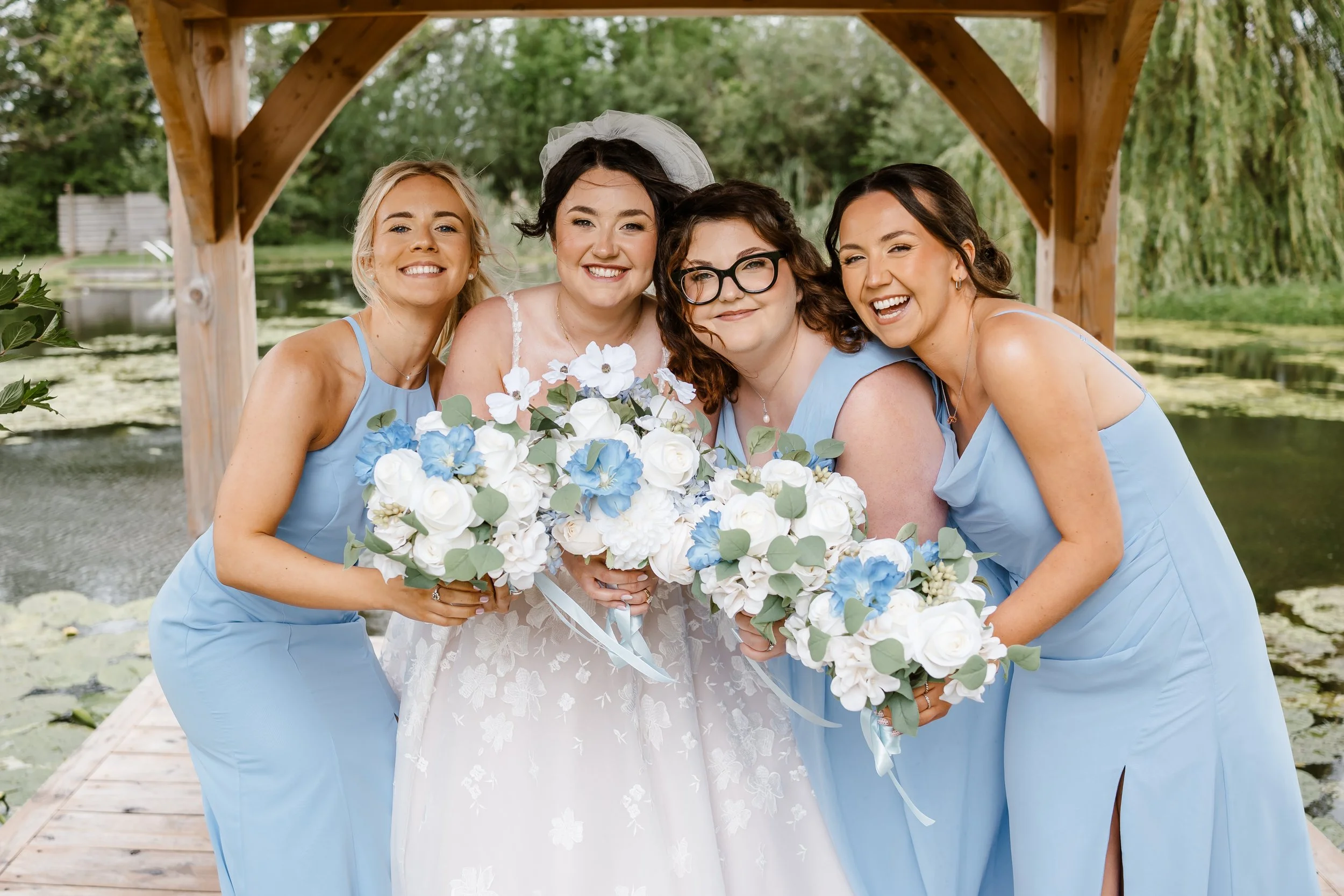 A bride and three bridesmaids smiling and hugging on a wooden dock by a pond. The bride is holding a bouquet of white and blue flowers. All women are dressed in light blue dresses, and the bride is wearing a white lace wedding gown and a veil. The ba