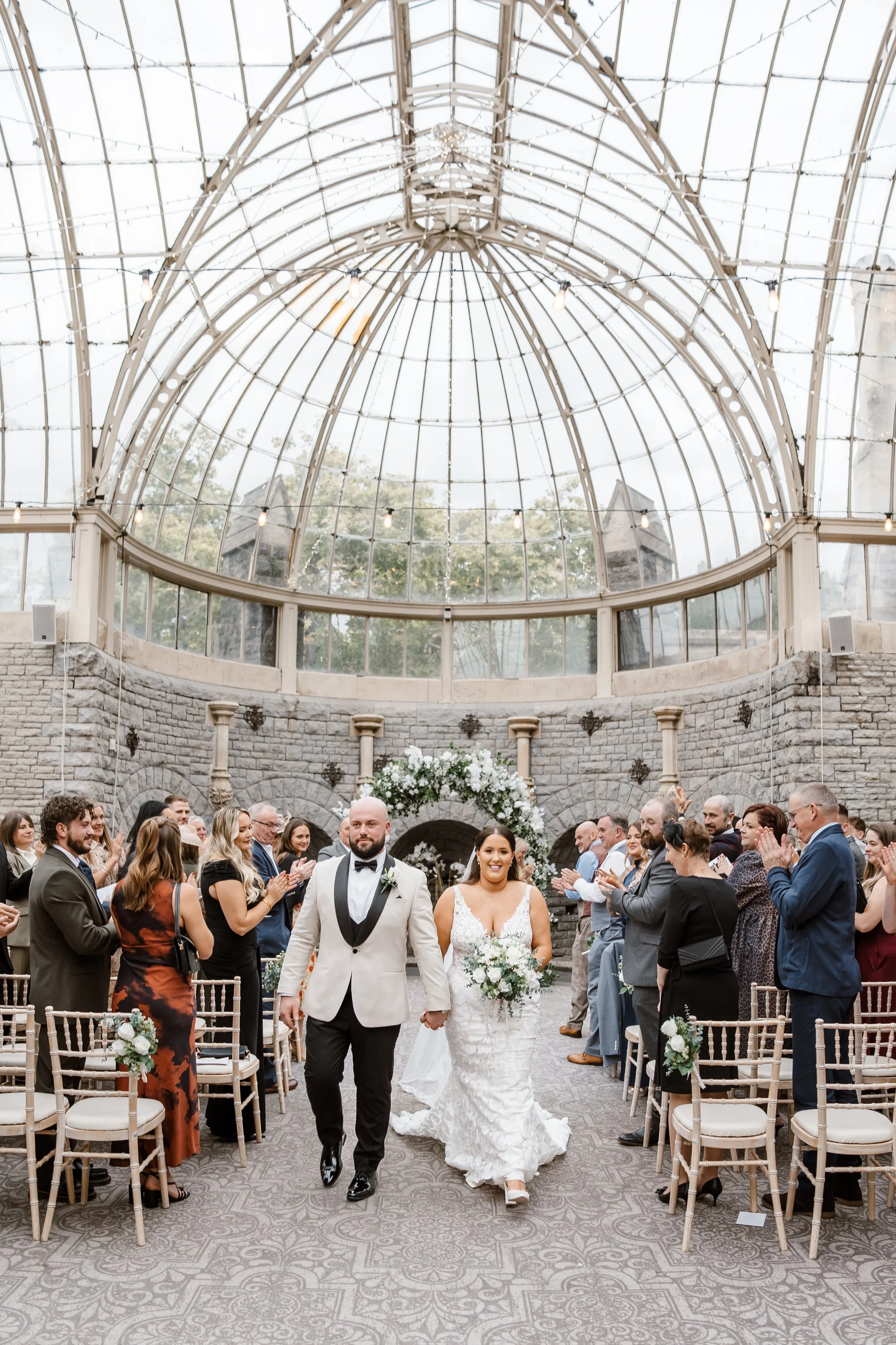 A bride and groom walking down the aisle at their wedding ceremony inside a glass-domed venue, surrounded by guests clapping and smiling.