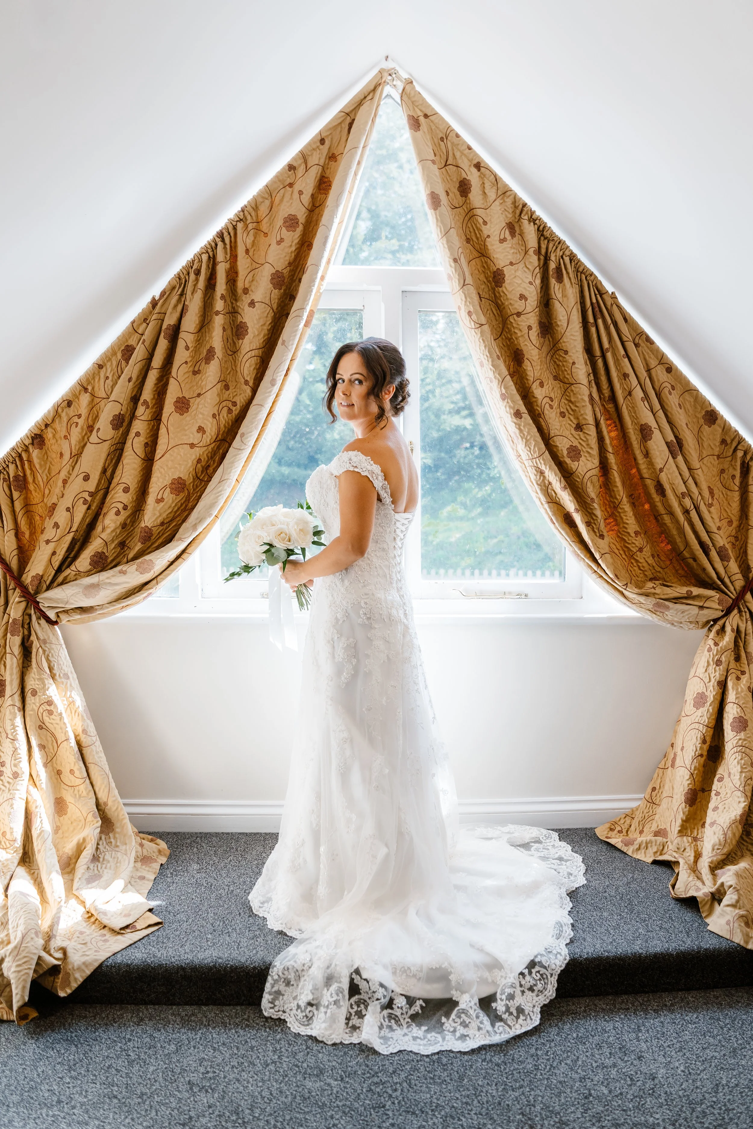 Bride in a white lace wedding gown holding a bouquet of white roses, standing in front of a window with gold patterned curtains tied back.