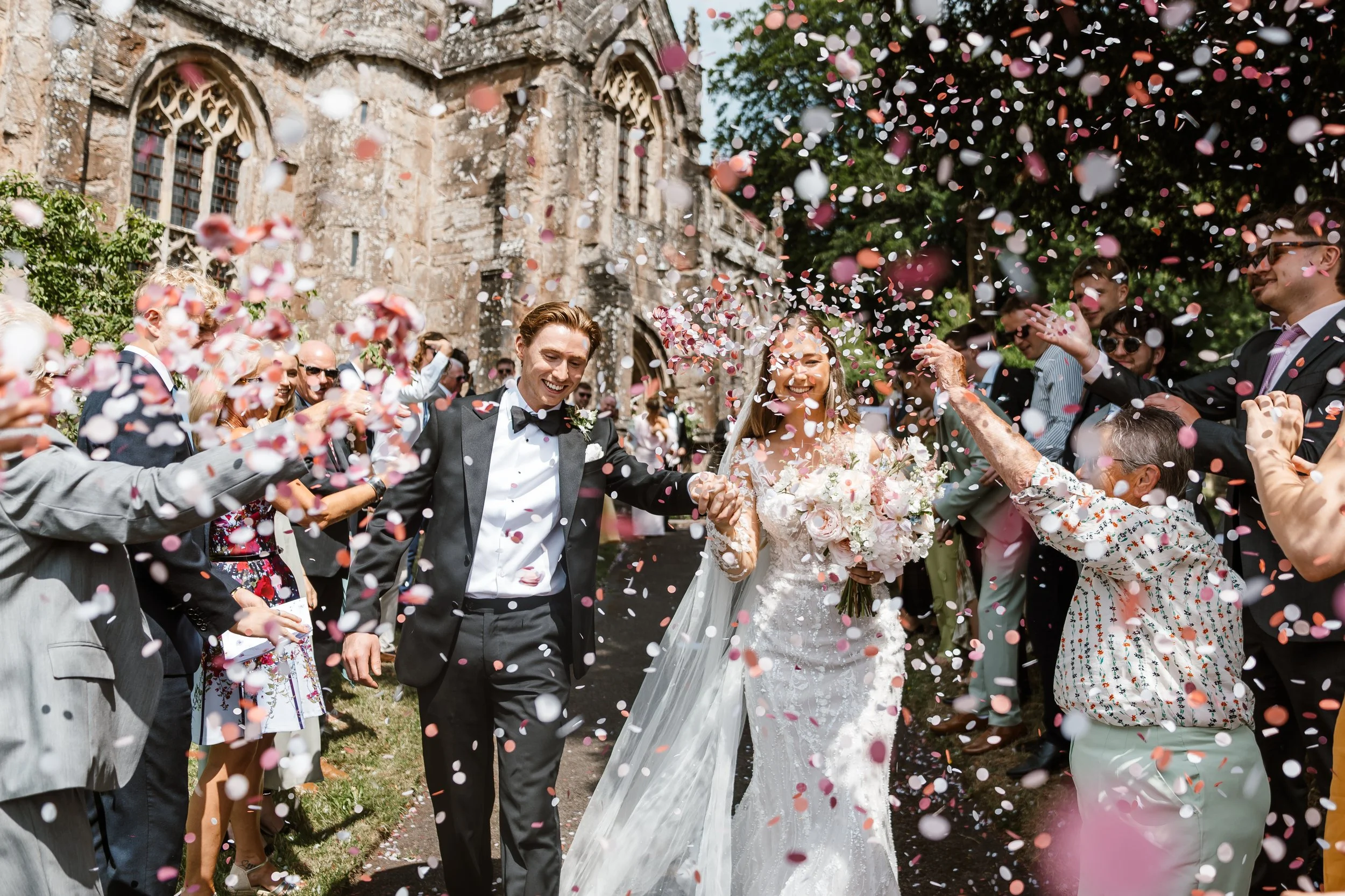 A newlywed couple walking hand in hand, surrounded by guests throwing confetti outside a church.