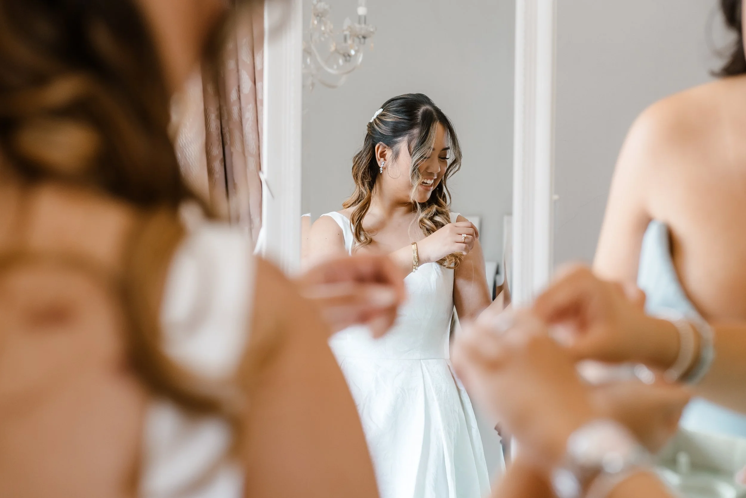 A bride dressed in white is smiling while getting ready, with other women around her also preparing, in a room with elegant decor and a chandelier.