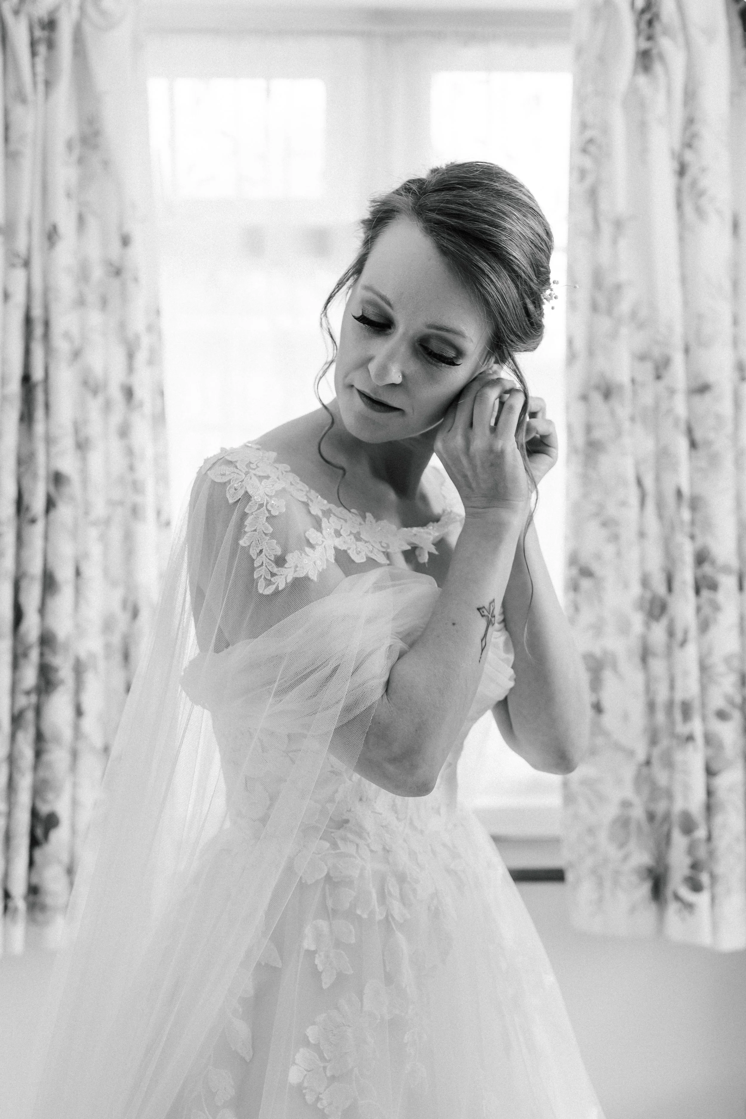 Black and white image of a woman in a wedding dress with lace details, touching her ear, with floral curtains and window in background.