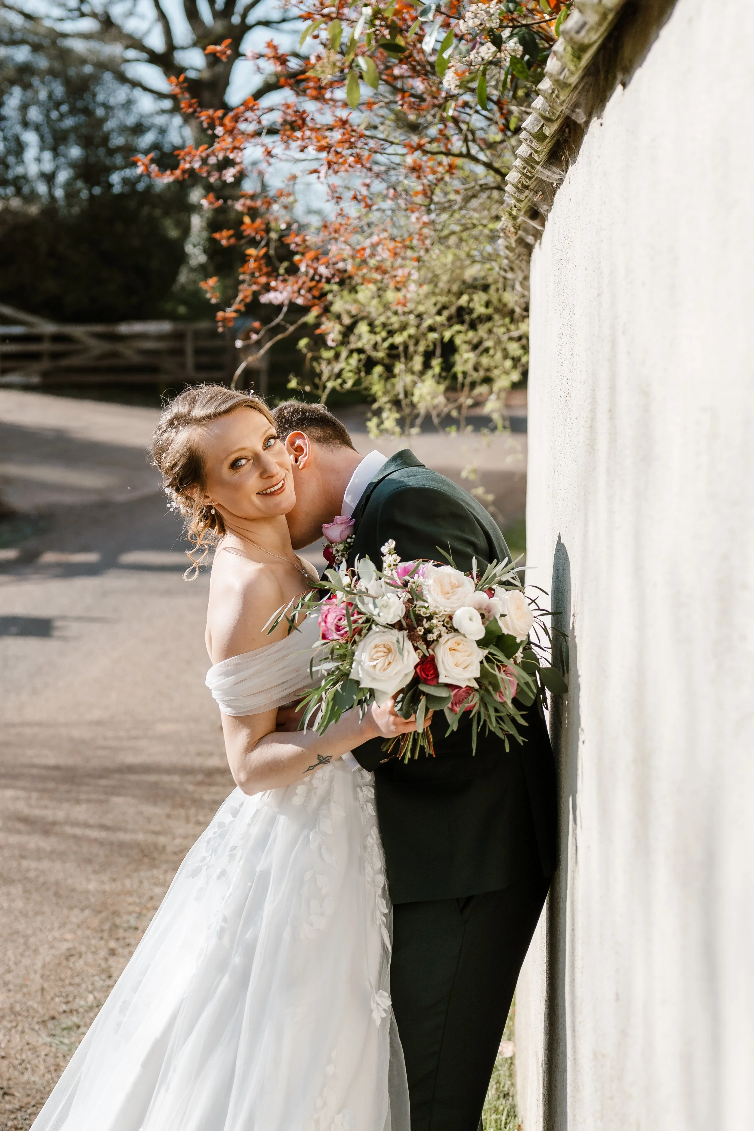 A bride and groom share a kiss outdoors, with the groom leaning against a white wall and the bride holding a bouquet of flowers, under blooming trees.