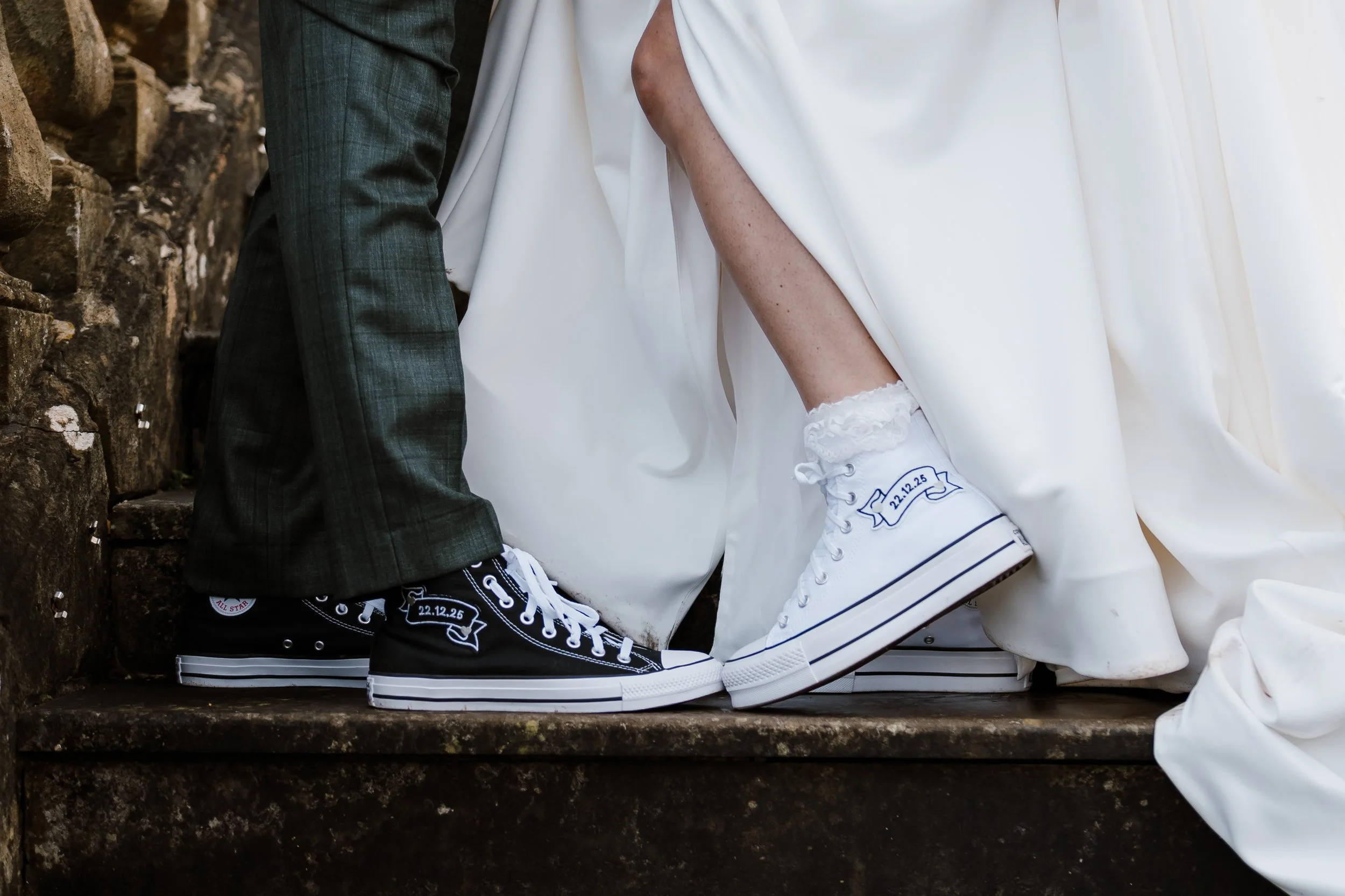 Close-up of the shoes and lower attire of a couple on their wedding day, with the bride wearing white sneakers with a date embroidered on them and the groom wearing black sneakers, both standing on stone steps.
