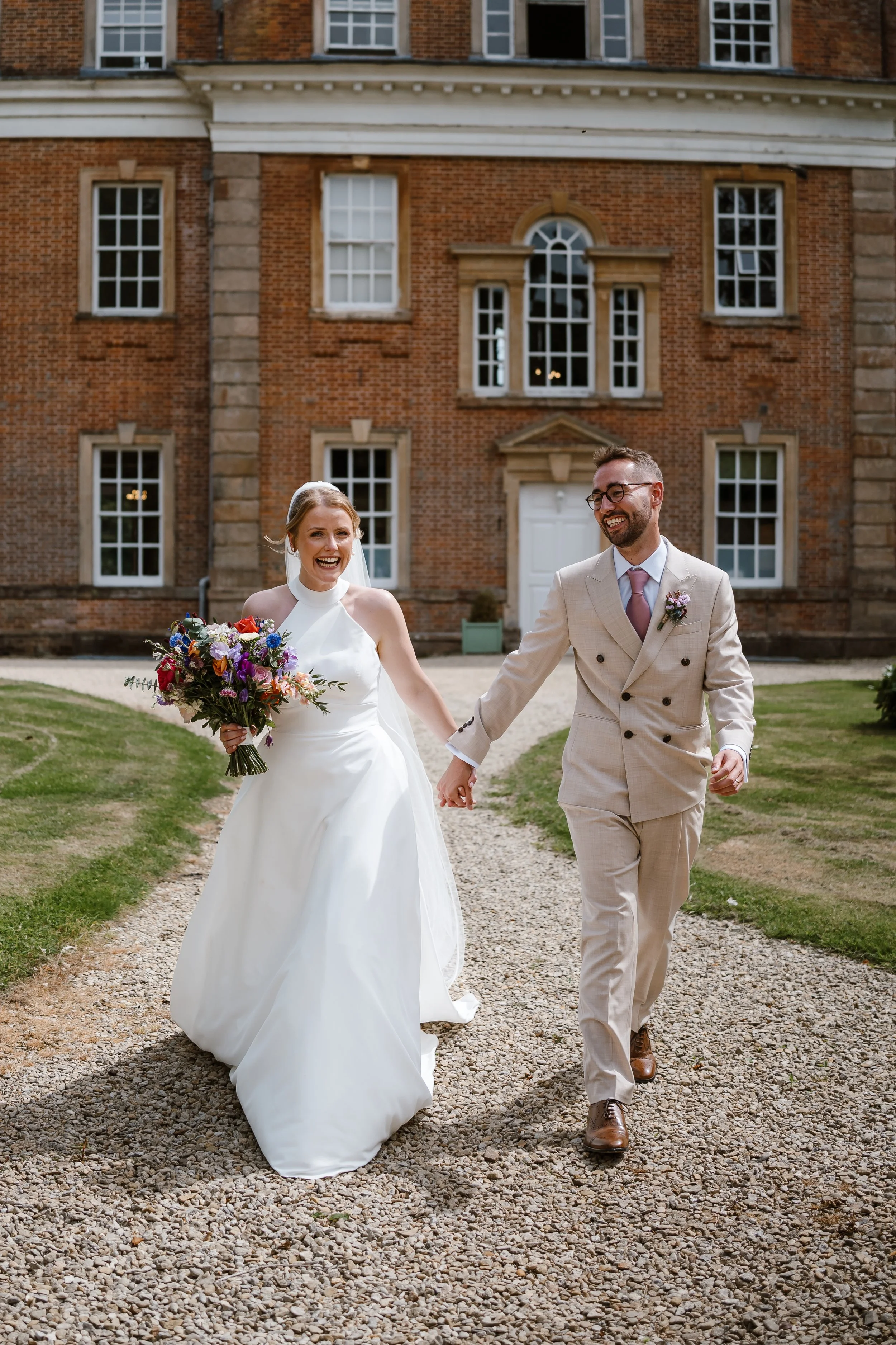A bride and groom walking hand in hand outside a brick building with multiple windows, smiling and looking happy.