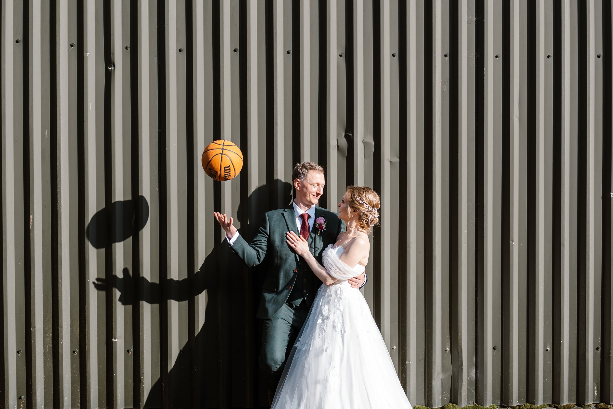 A newly married couple dressed in wedding attire standing close and looking at each other, smiling, with a tall, vertical striped gray wall in the background; a basketball is floating in mid-air near the top of the image.